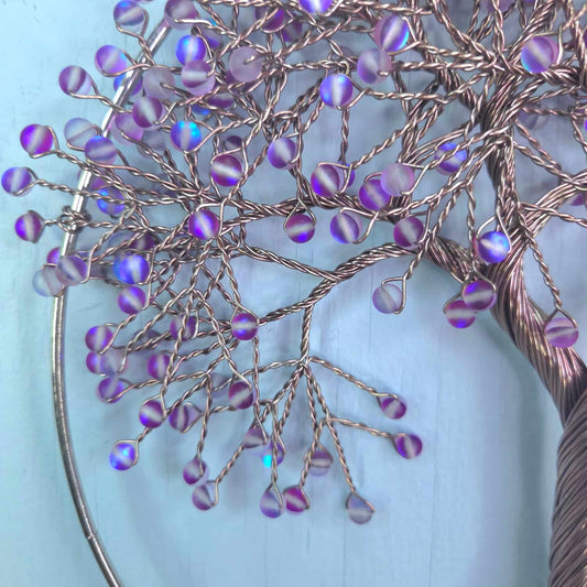 A close-up of a decorative wire tree branch arrangement with iridescent purple and blue bead-like accents, set against a light blue background.