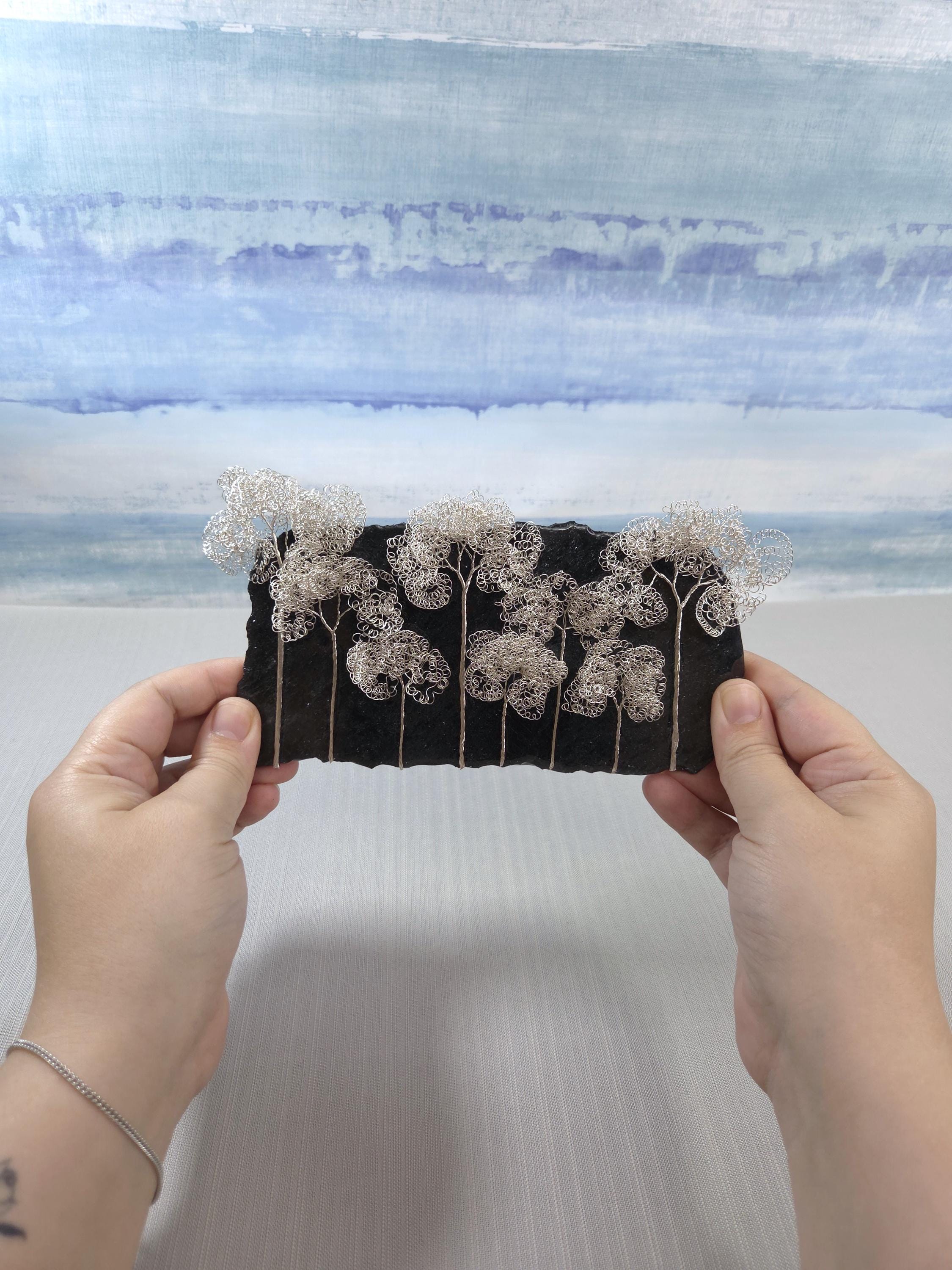 Handmade jewelry piece held between two hands with a watercolor landscape in the background