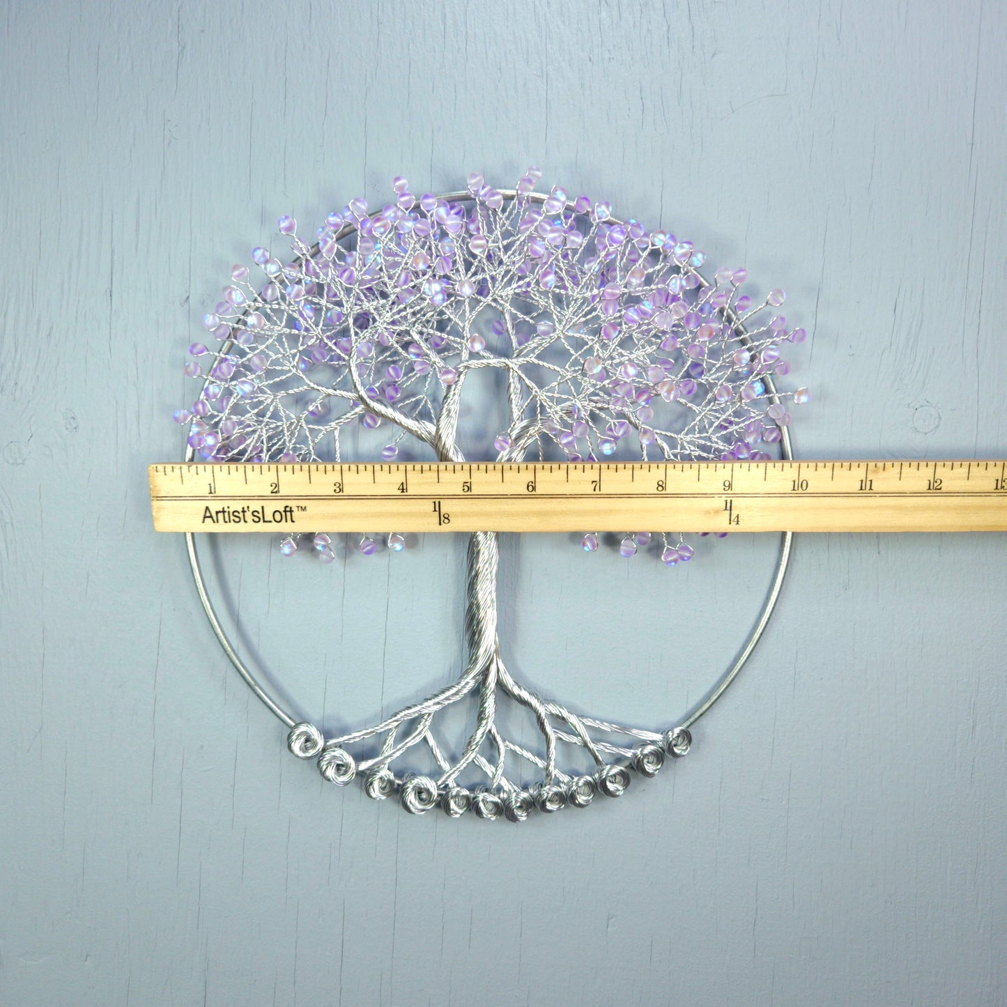 Decorative wall art of a tree with purple flowers and silver base, measured against a ruler on a light gray background.
