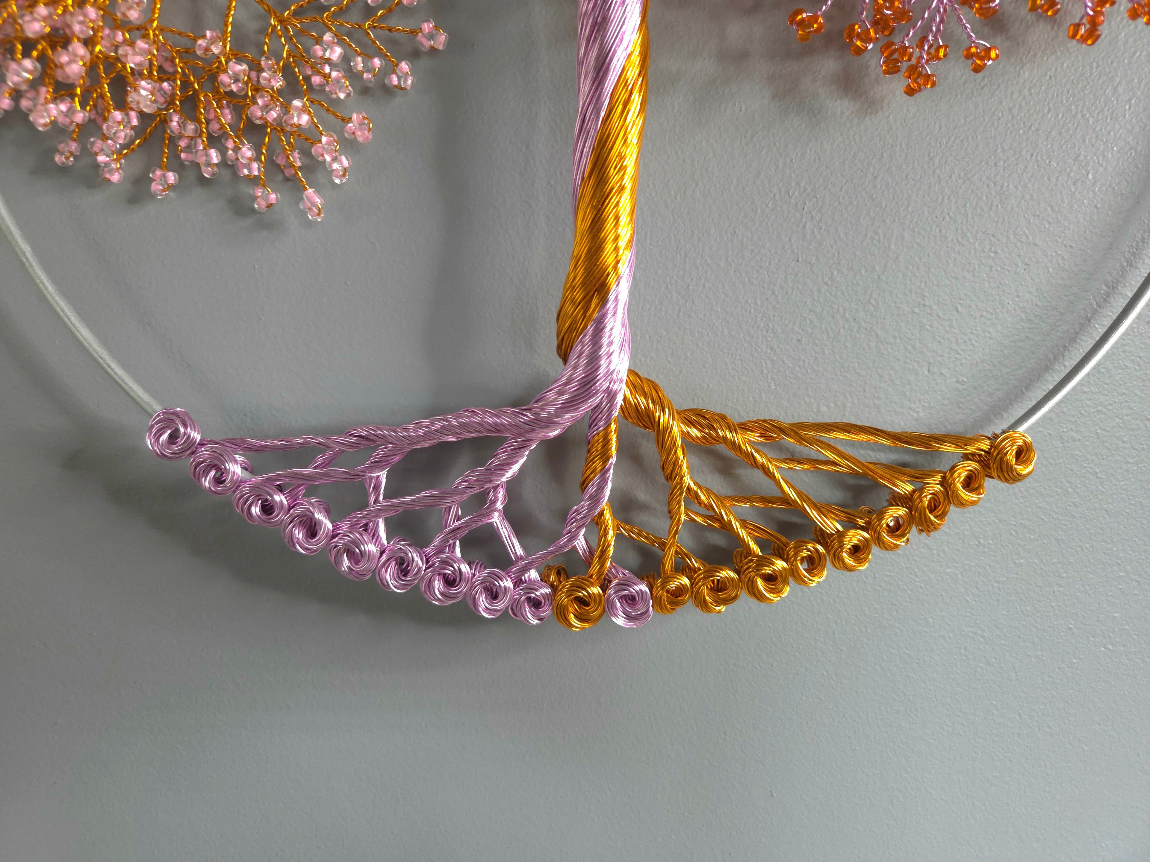 Close-up of a decorative wreath with pink and gold beaded branches on a light gray background.