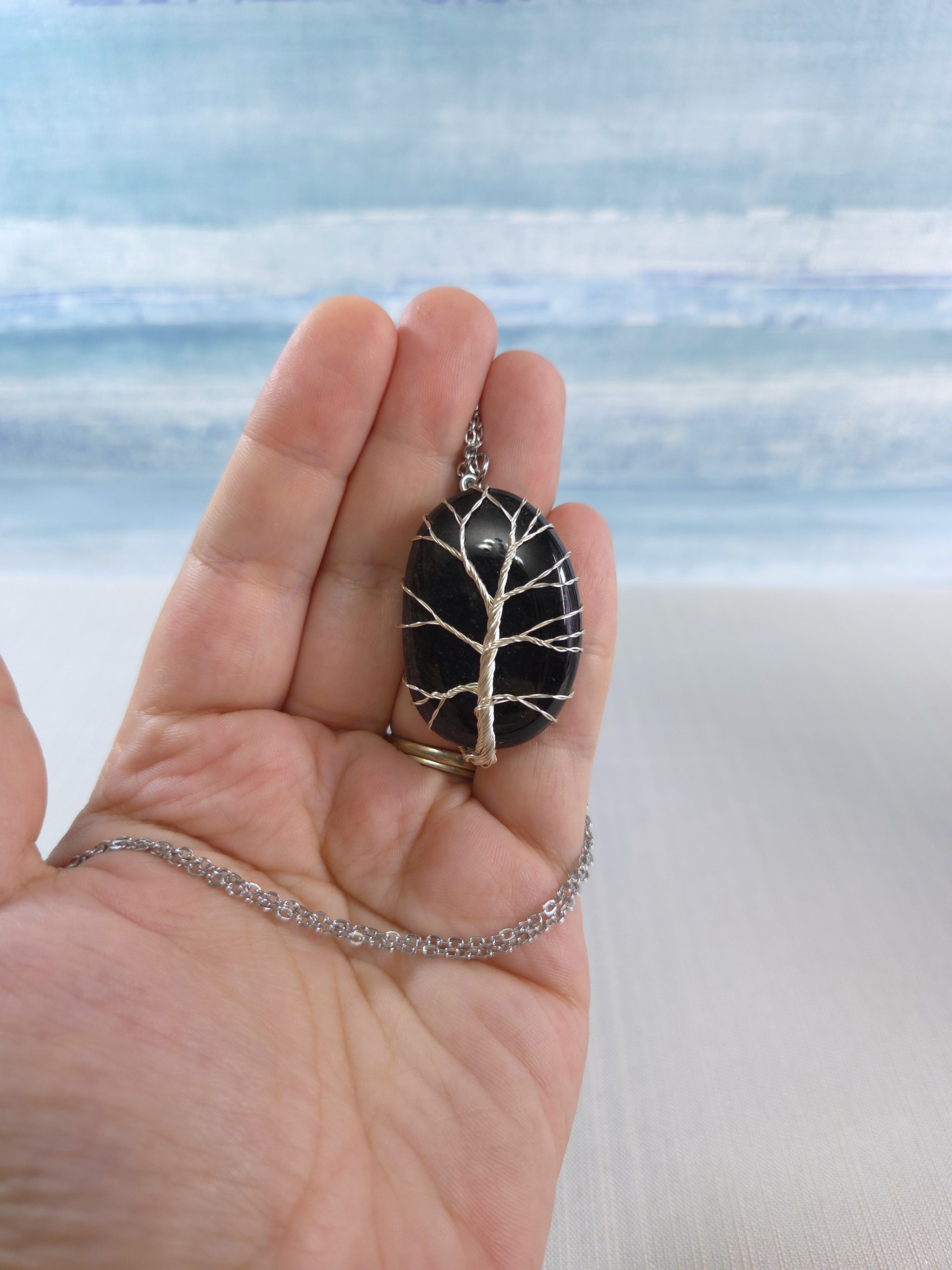 Hand holding a necklace with a tree-of-life pendant against a beach background