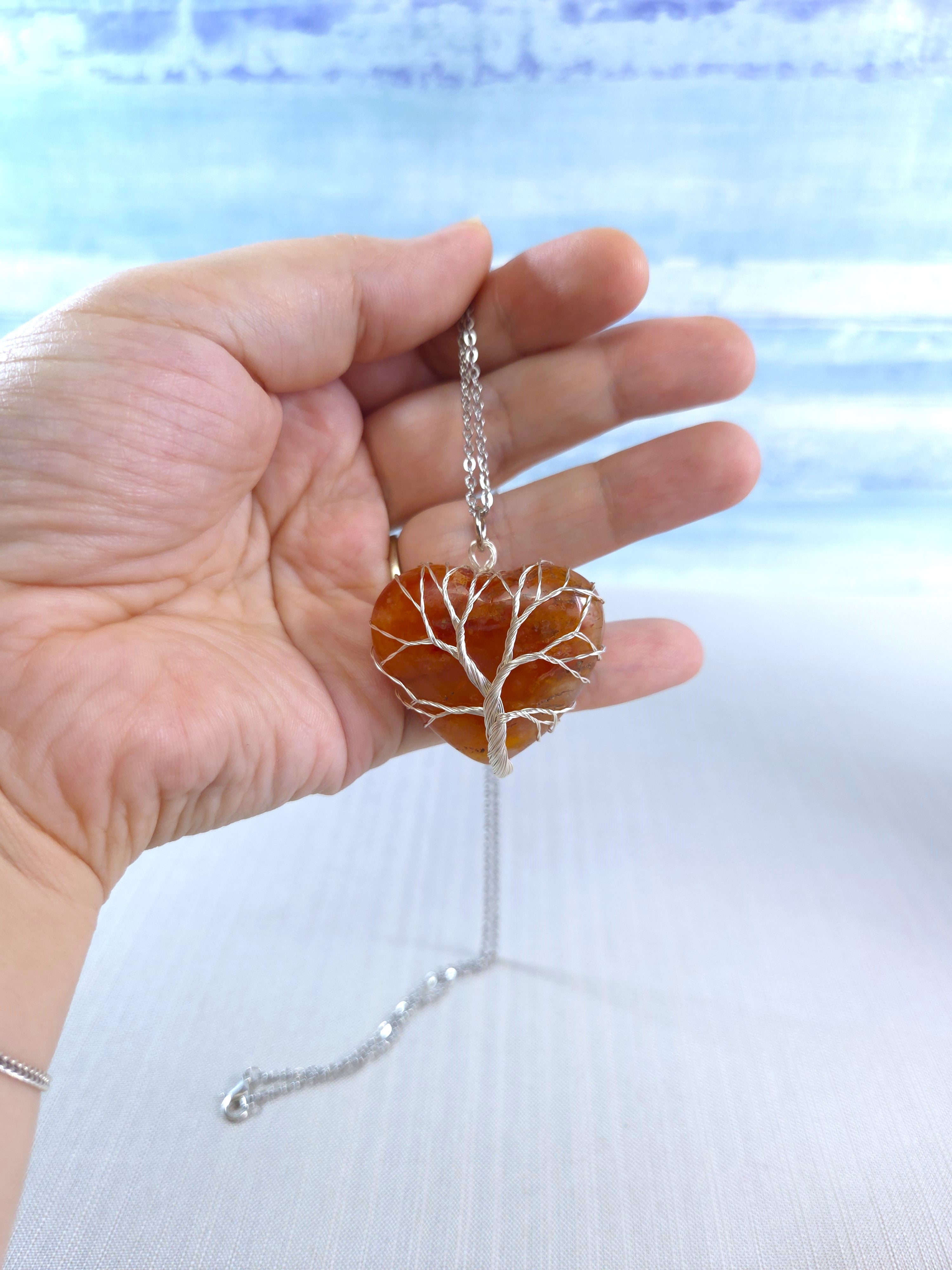 Hand holding a necklace with a tree of life pendant against a light blue background