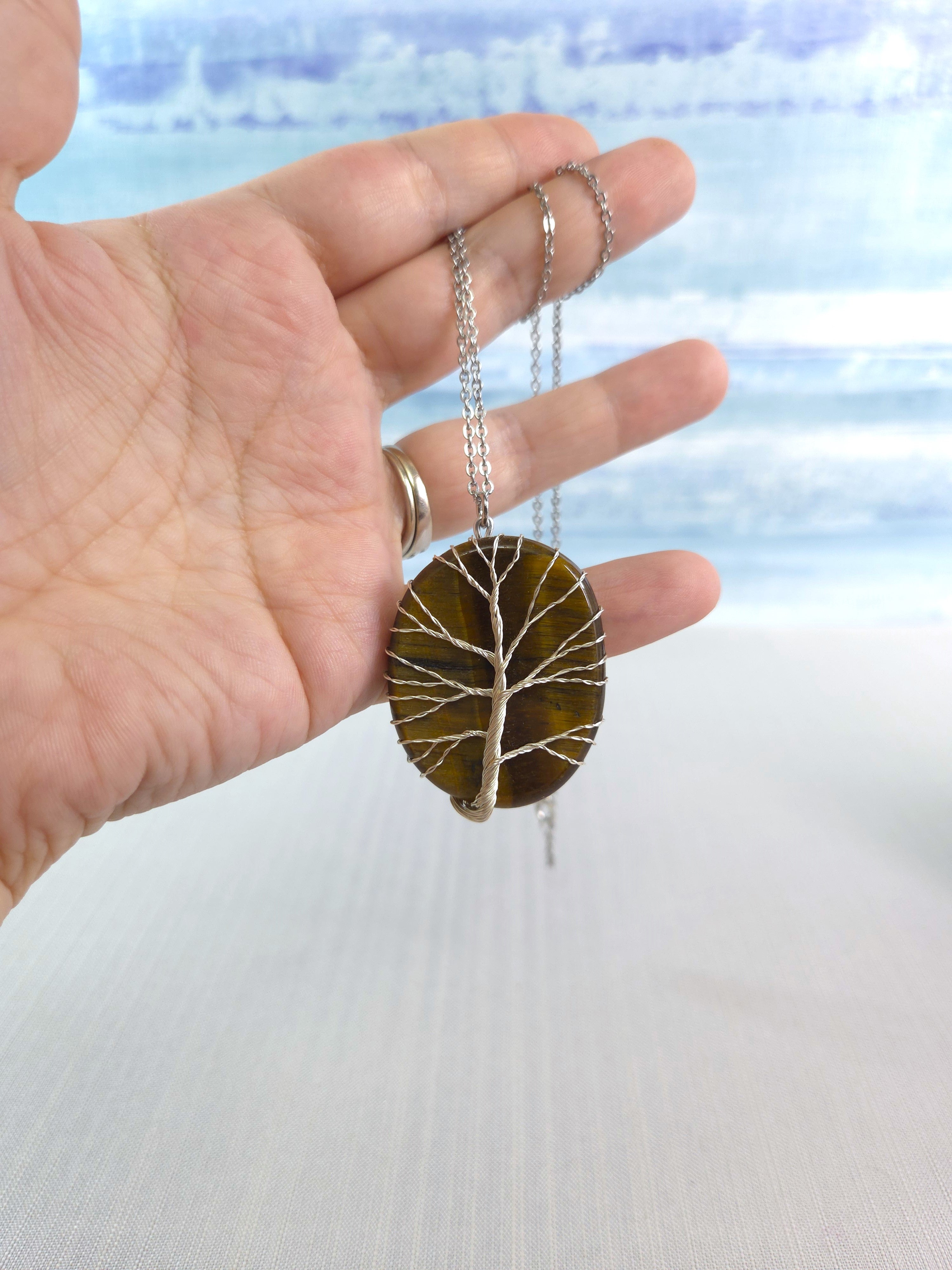 Hand holding a necklace with a tree-of-life pendant against a blurred natural background