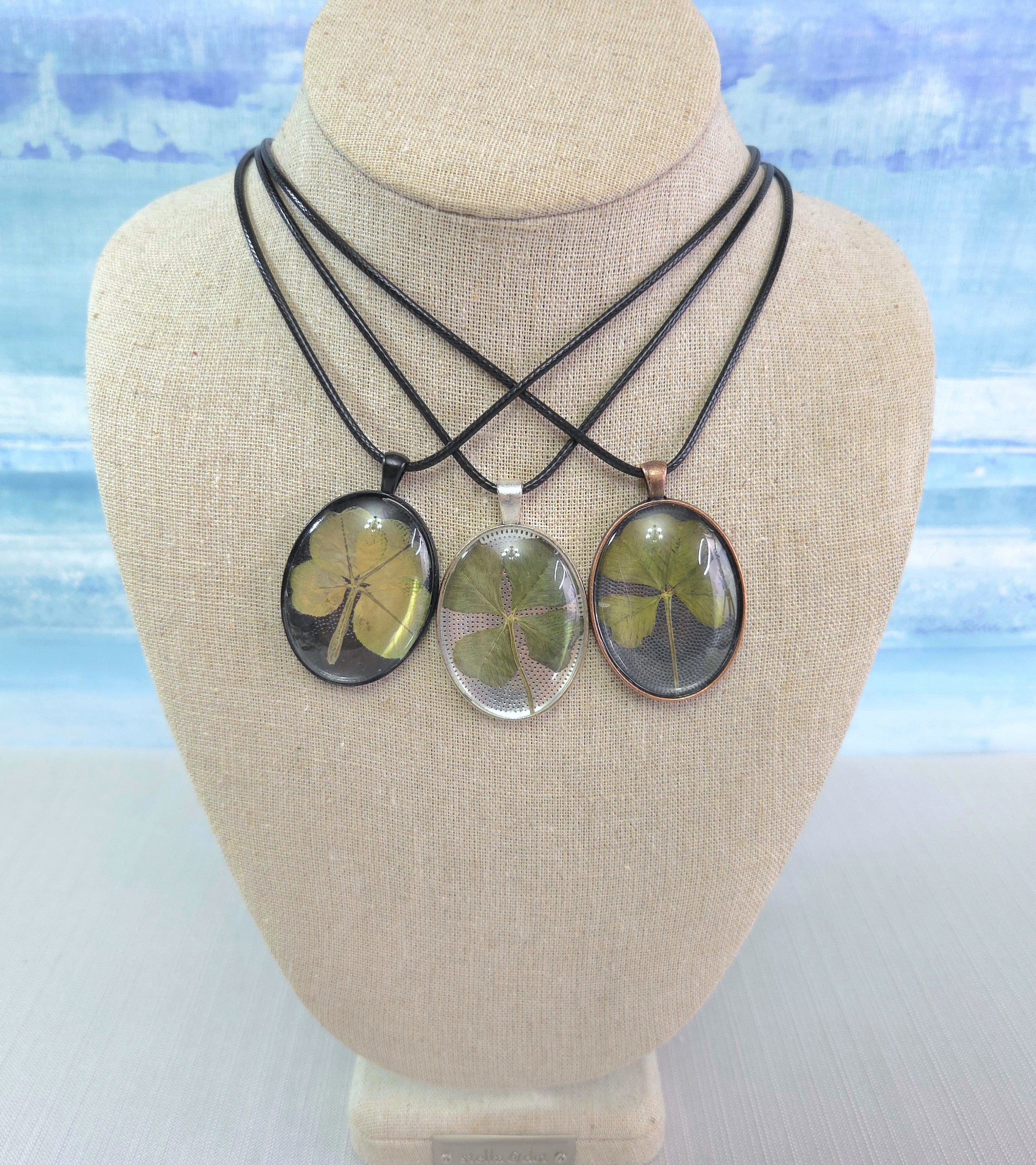 Three necklaces with oval pendants on a mannequin bust against a beach-themed background.