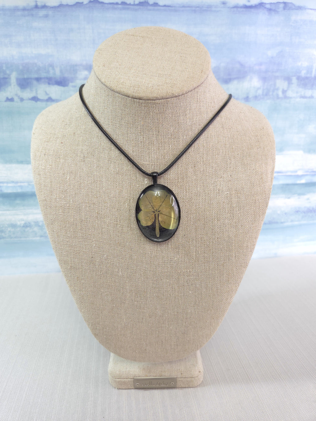 Necklace with a clover pendant on a beige bust against a light blue watercolor background