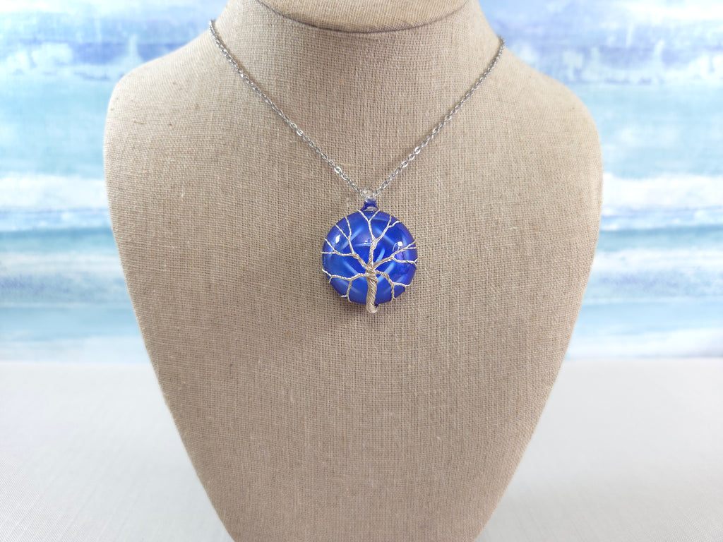 Blue tree-shaped pendant necklace on a mannequin bust with a blurred beach background