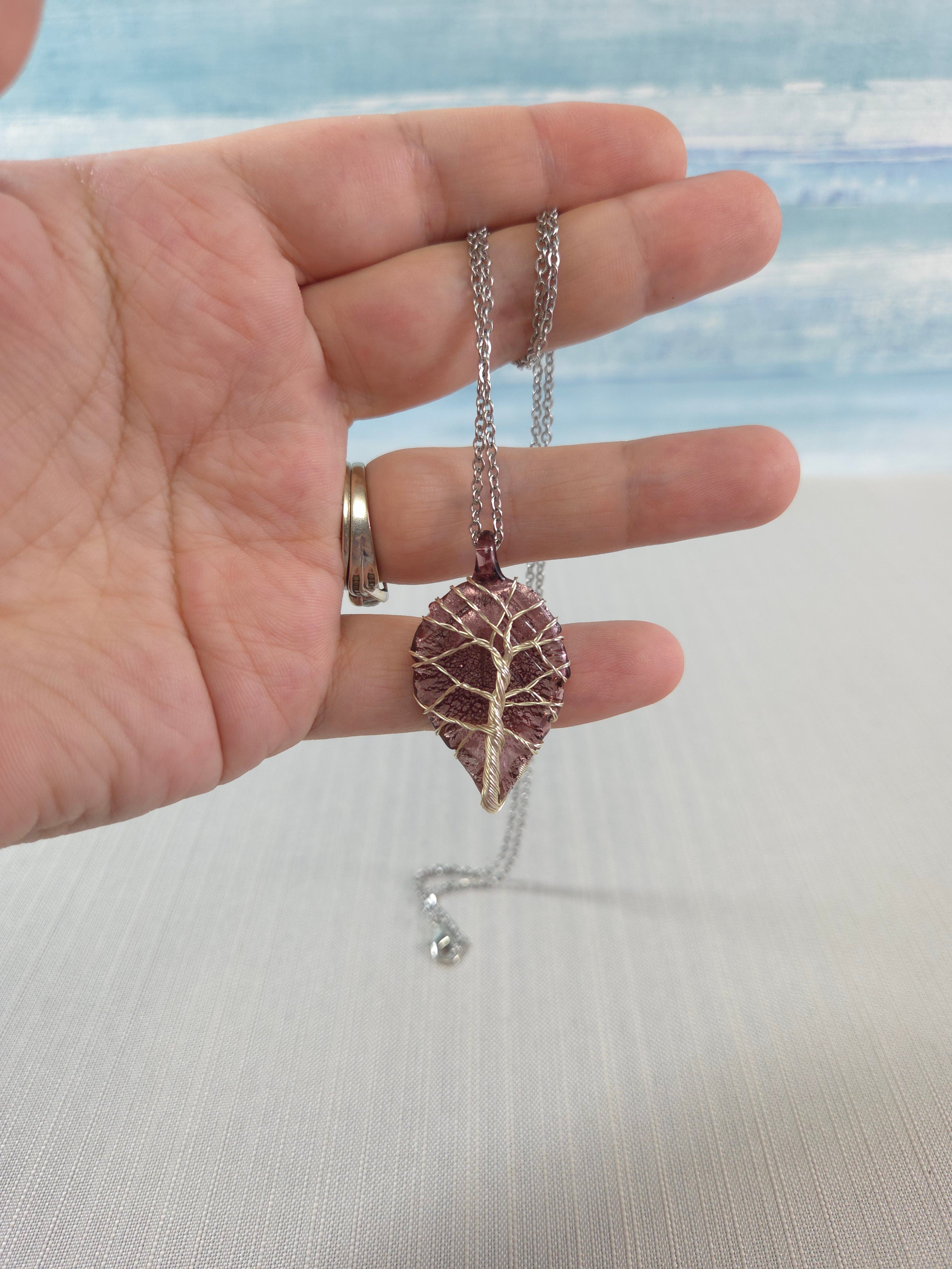 Hand wearing a leaf-shaped ring with a necklace on a neutral background