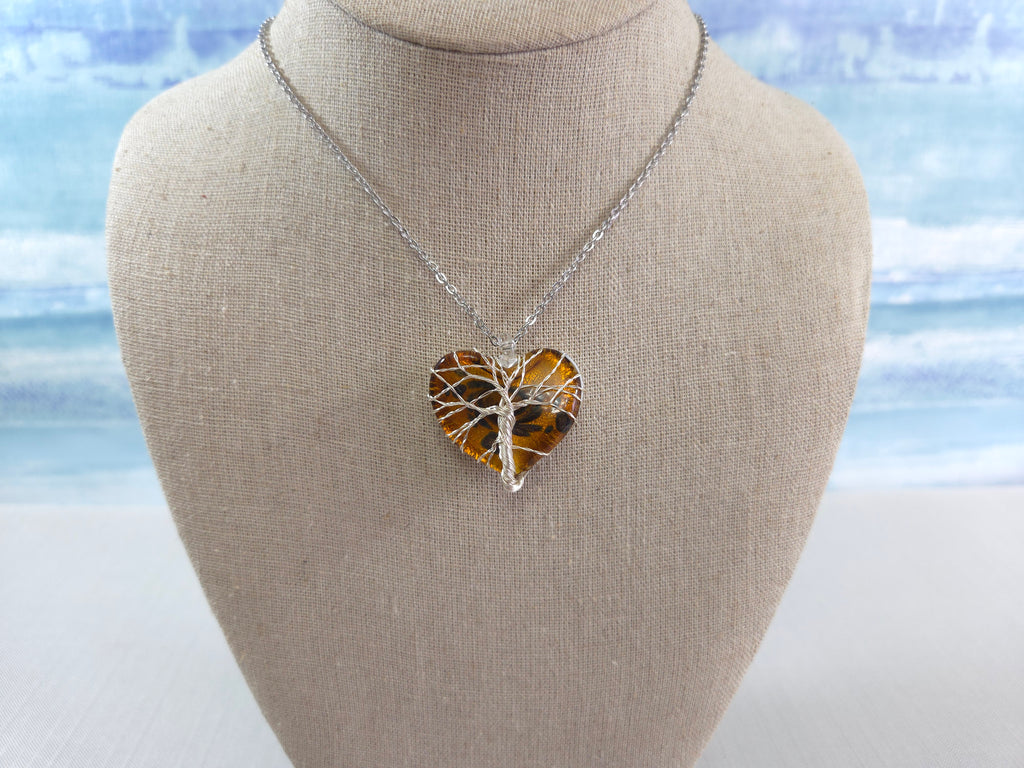 Heart-shaped pendant with tree design on a necklace, displayed on a bust against a blurred natural background.