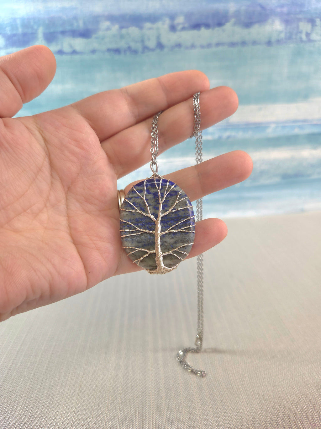 Hand holding a necklace with a tree-of-life pendant against a blurred beach background