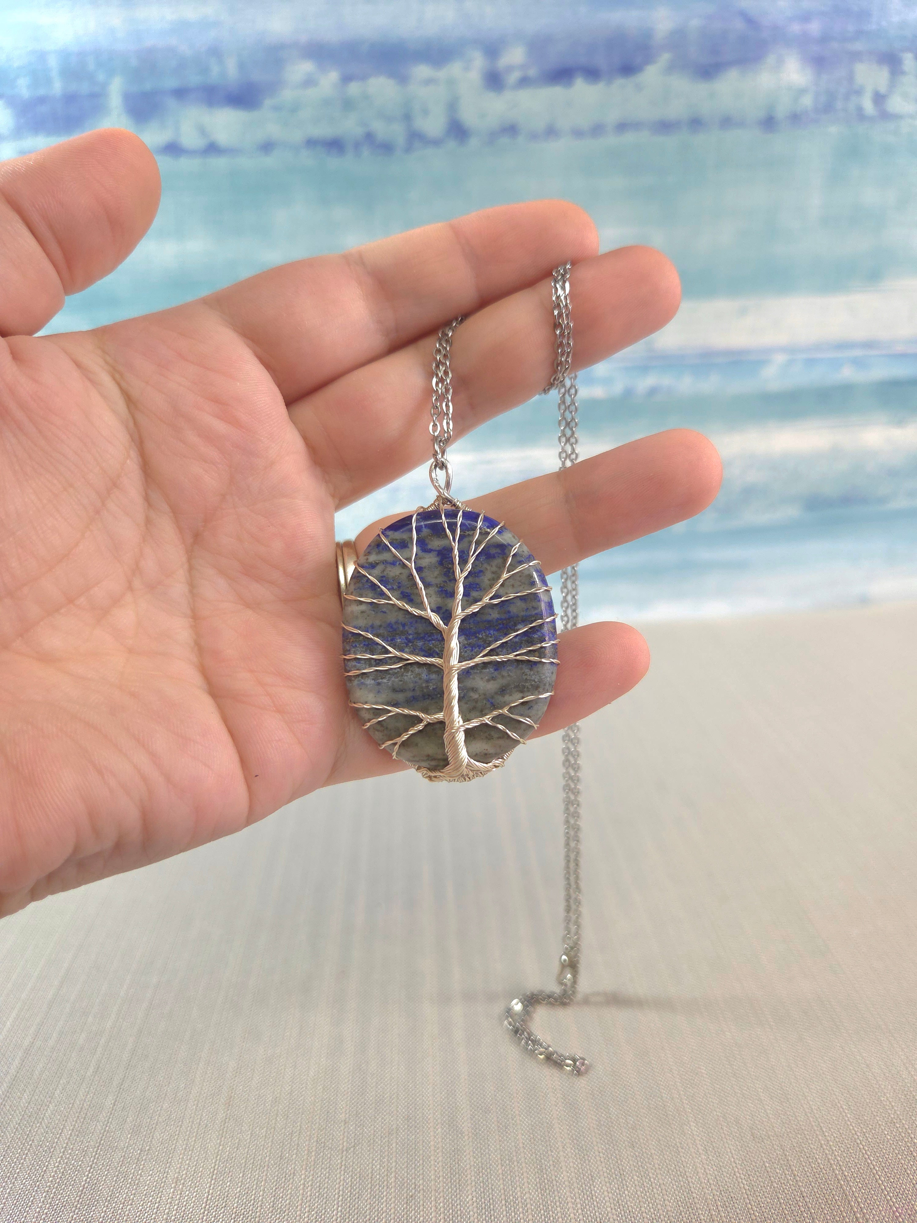 Hand holding a necklace with a tree-of-life pendant against a blurred beach background