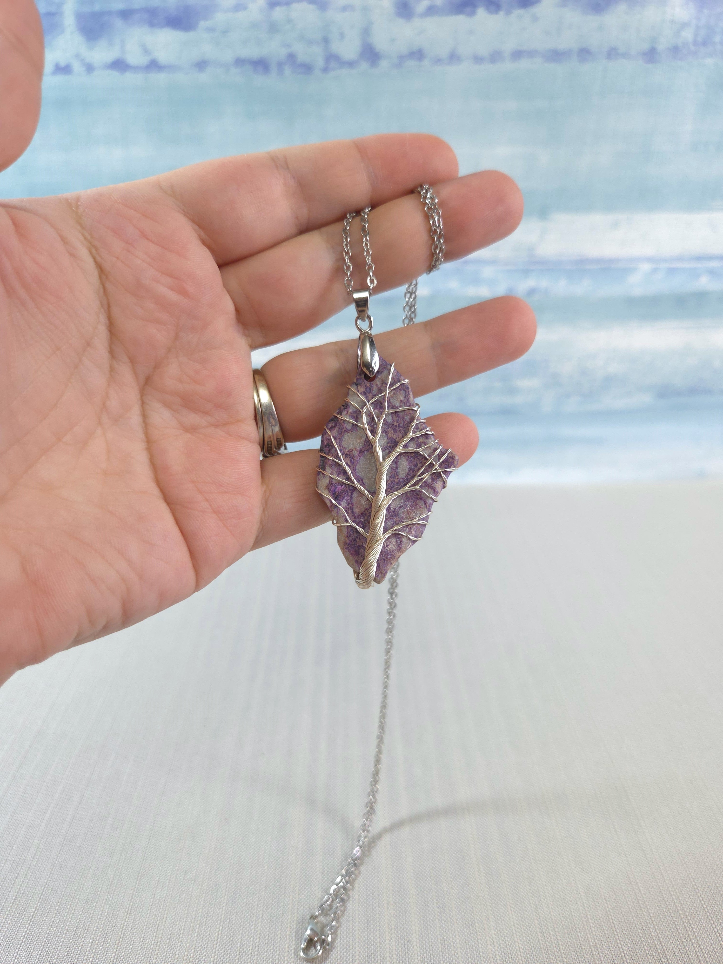 Hand holding a purple leaf-shaped pendant with a silver chain against a light blue and white background.