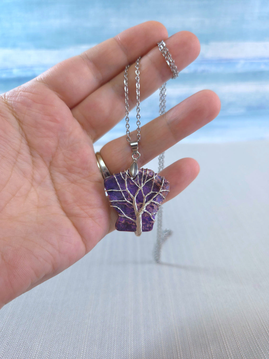 Hand holding a necklace with a purple tree pendant against a blurred beach background