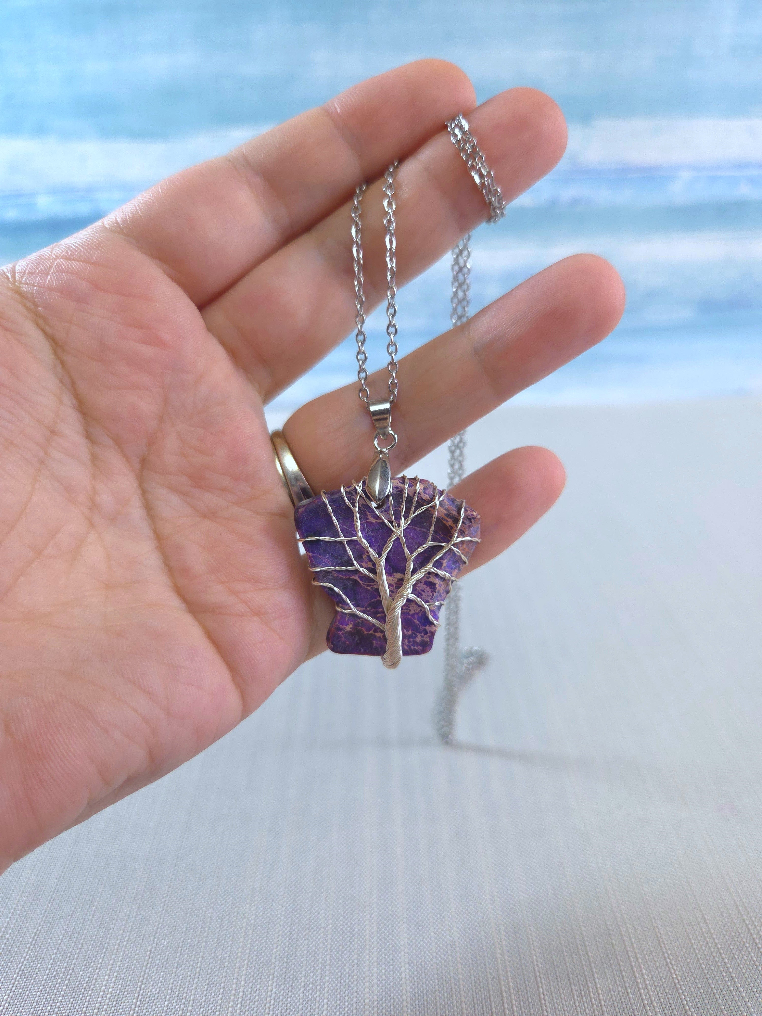 Hand holding a necklace with a purple tree pendant against a blurred beach background