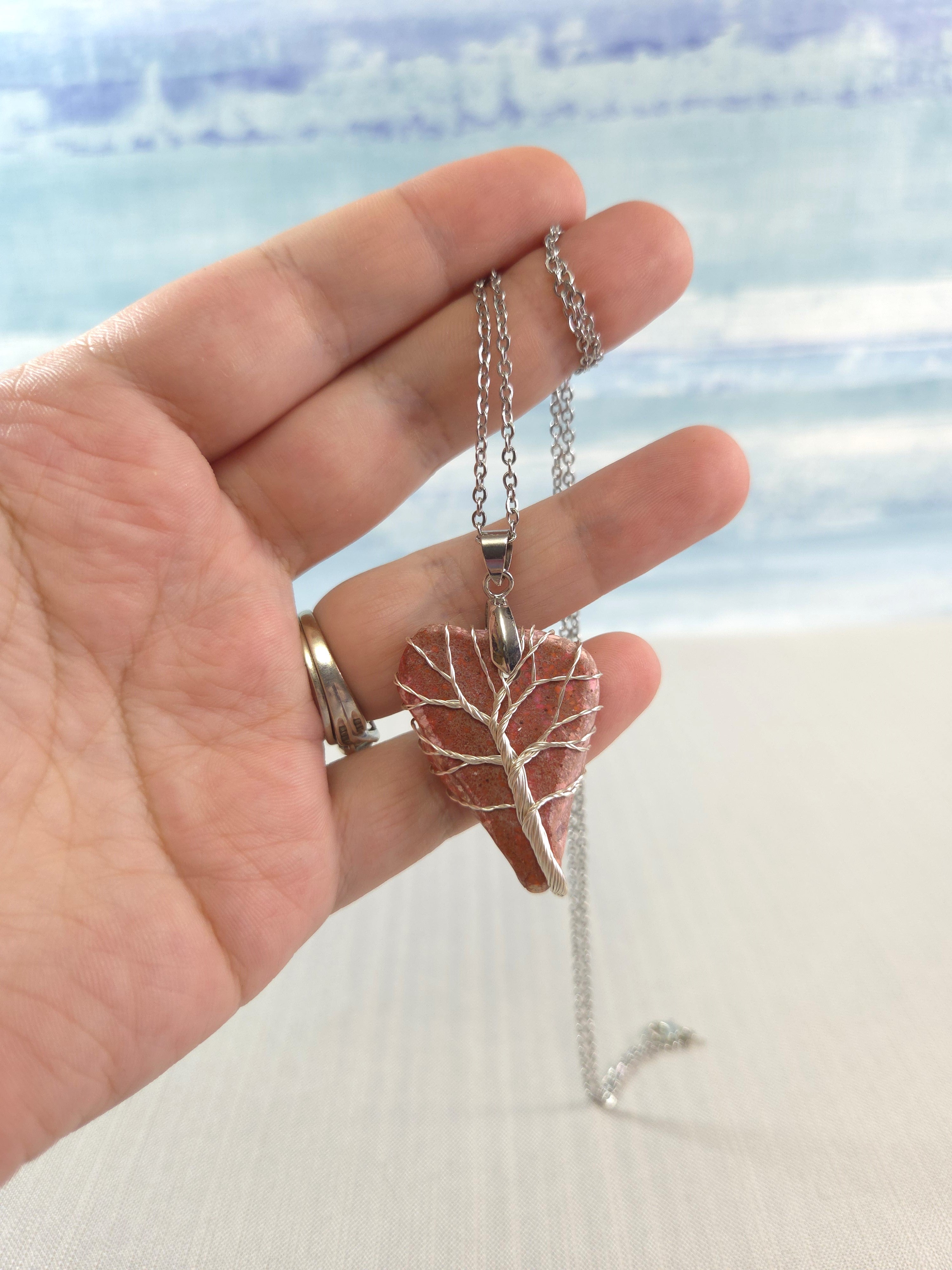Hand holding a necklace with a heart-shaped pendant against a blurred beach background