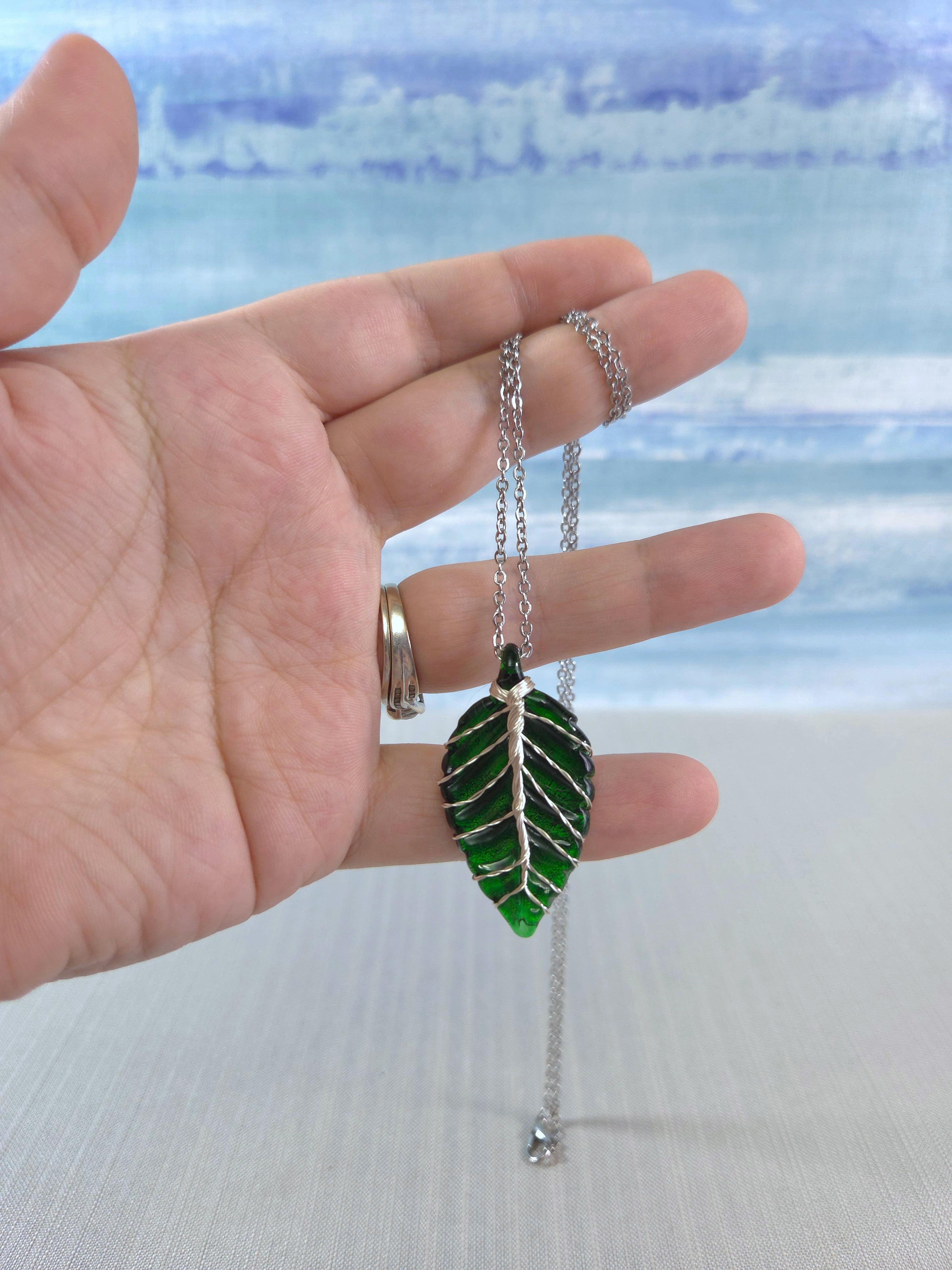 Hand holding a green leaf-shaped pendant with a chain against a blurred natural background