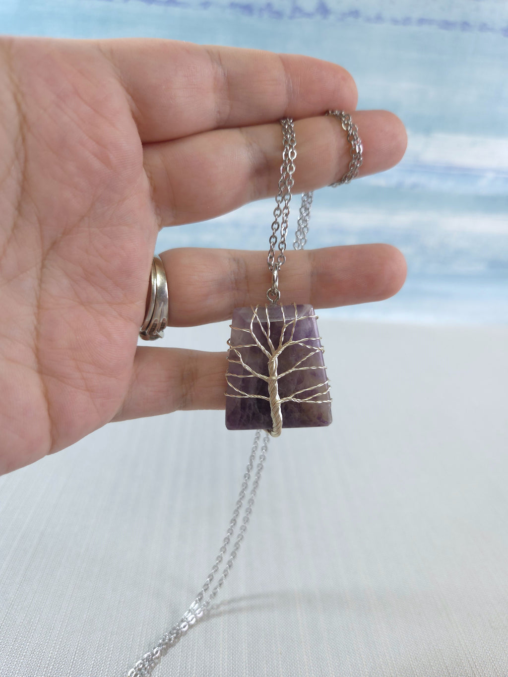 Hand holding a necklace with a purple stone and silver chain against a blurred natural background