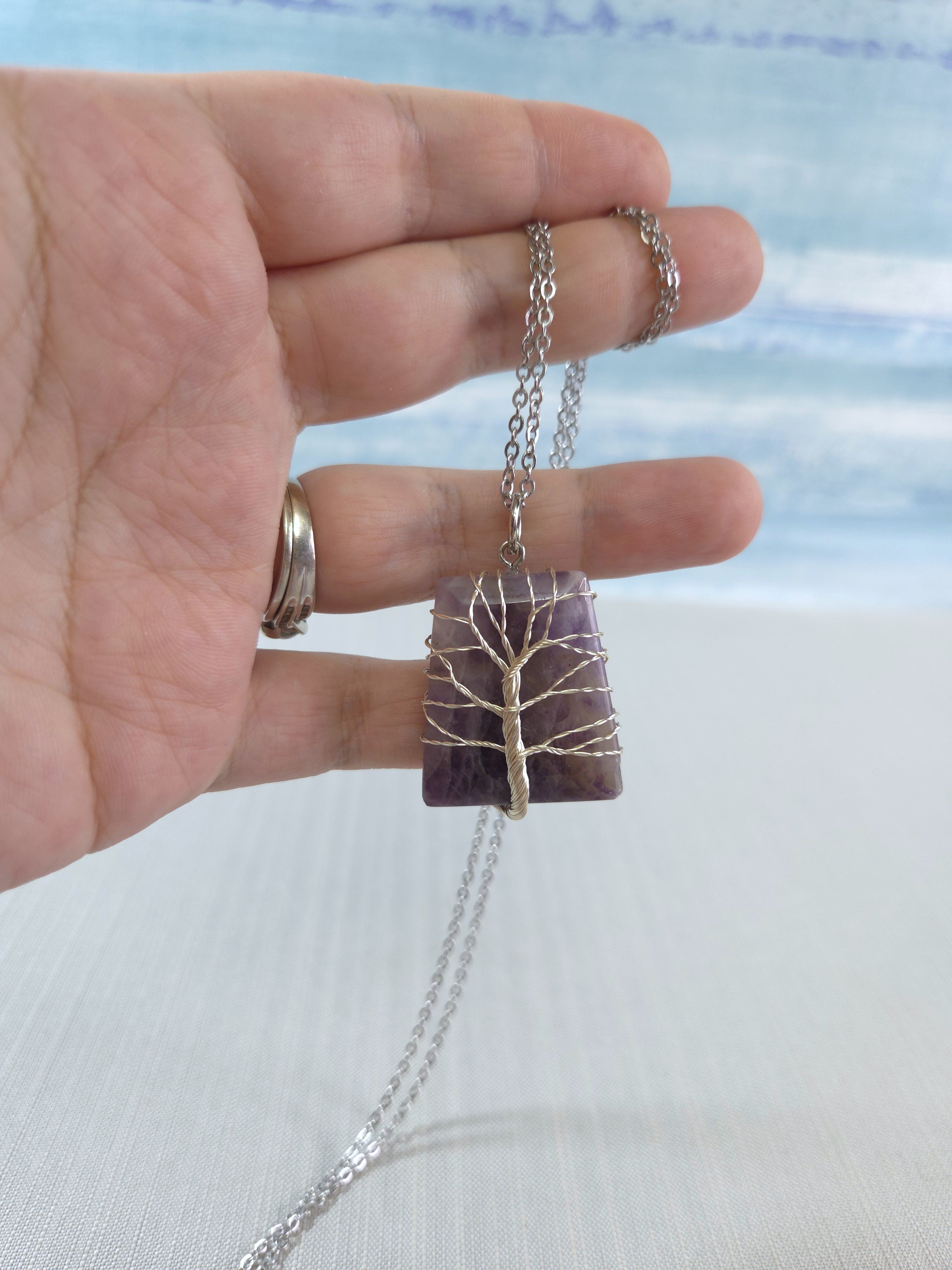 Hand holding a necklace with a purple stone and silver chain against a blurred natural background