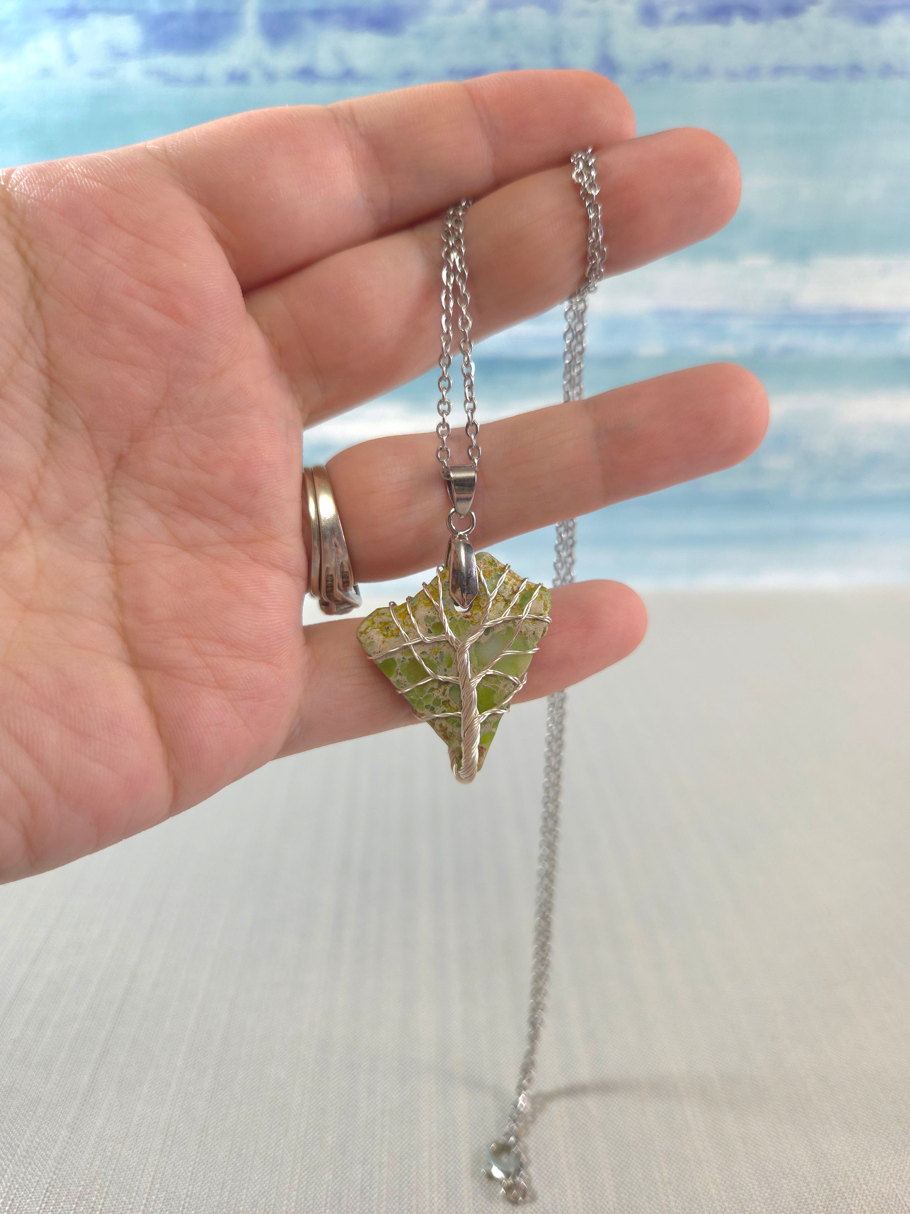 Hand holding a silver necklace with a leaf pendant against a beach background