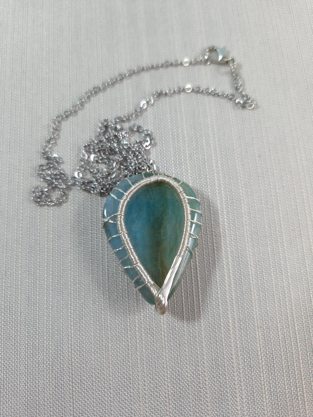 Teardrop-shaped pendant with blue stone on a silver chain against a light gray background