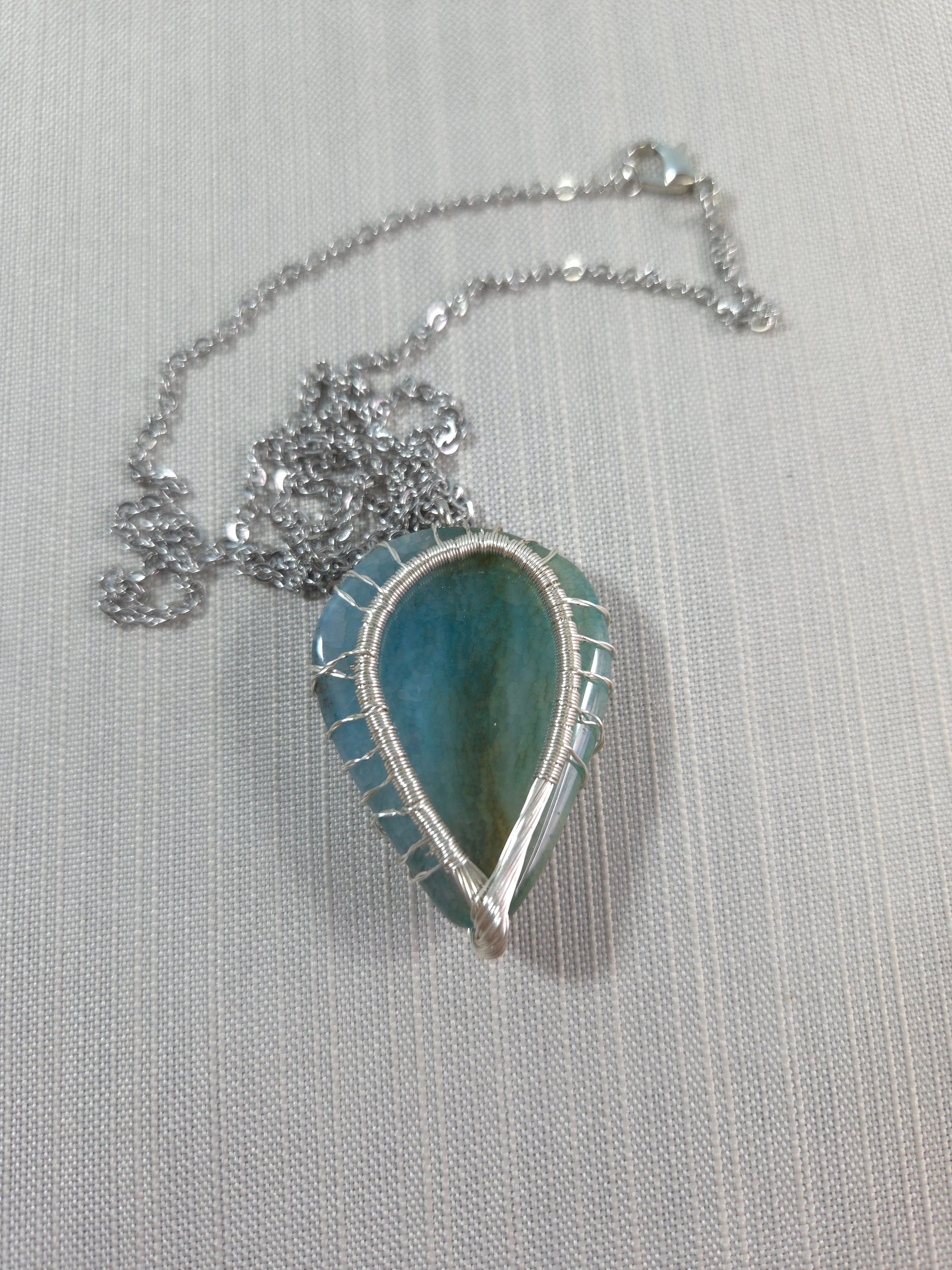 Teardrop-shaped pendant with blue stone on a silver chain against a light gray background