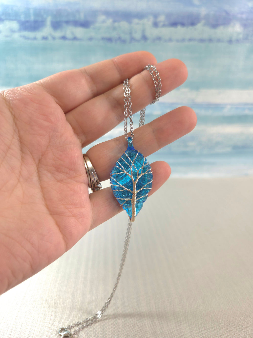 Hand holding a necklace with a blue leaf pendant against a blurred natural background