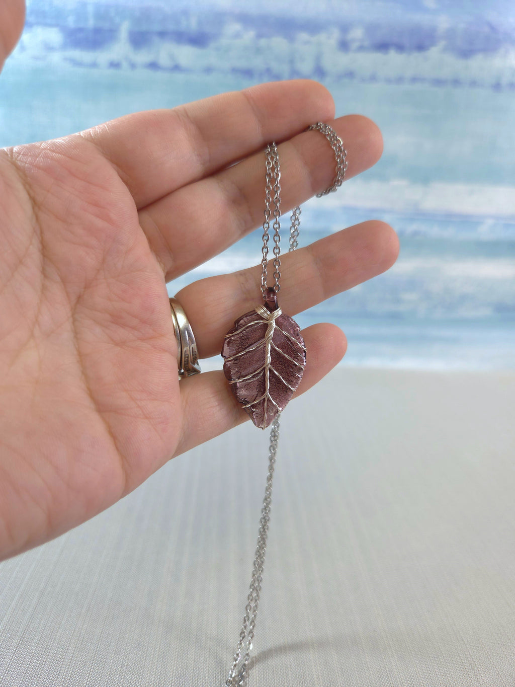 Hand holding a necklace with a leaf-shaped pendant against a beach background