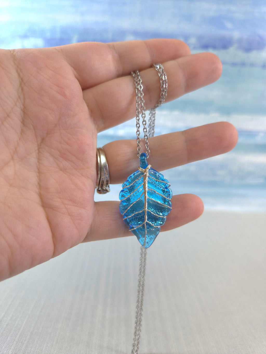 Hand holding a blue leaf-shaped pendant necklace with a blurred beach background
