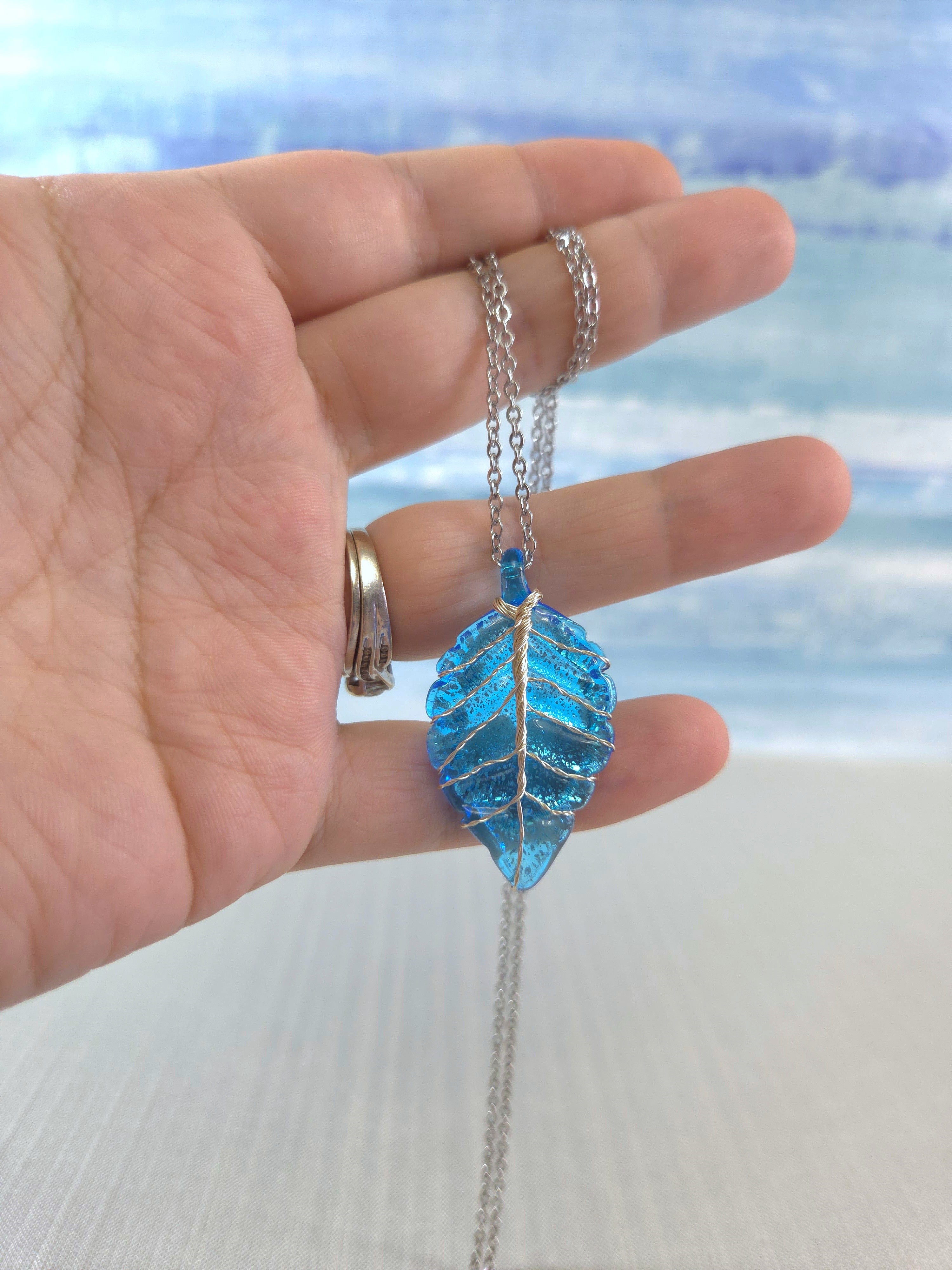 Hand holding a blue leaf-shaped pendant necklace with a blurred beach background