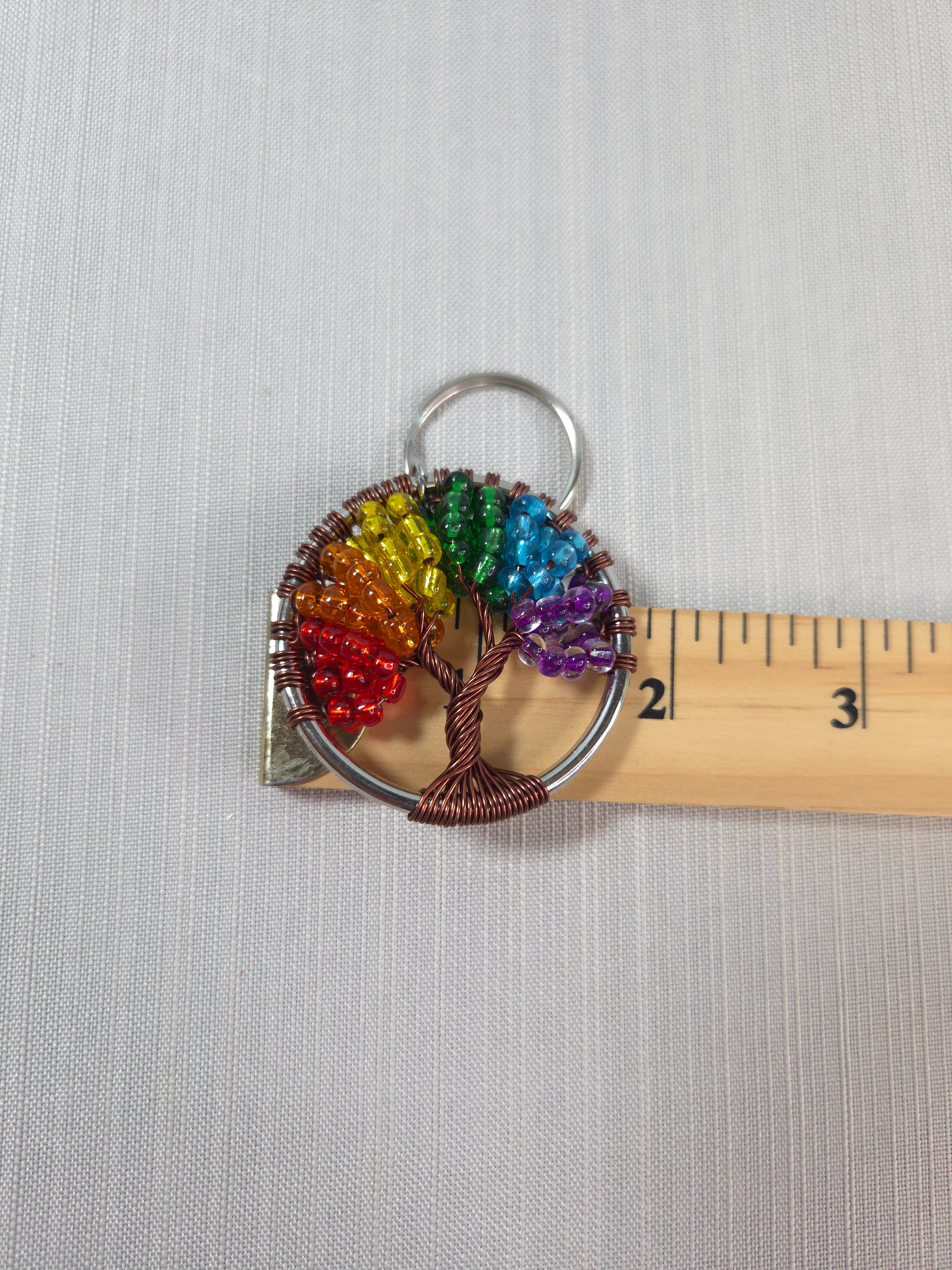 Rainbow-colored tree of life keychain with a ruler for scale on a gray background