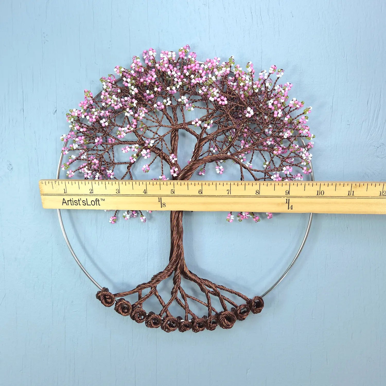 Decorative wall art of a tree with pink flowers and brown branches, measured against a ruler on a light blue background.
