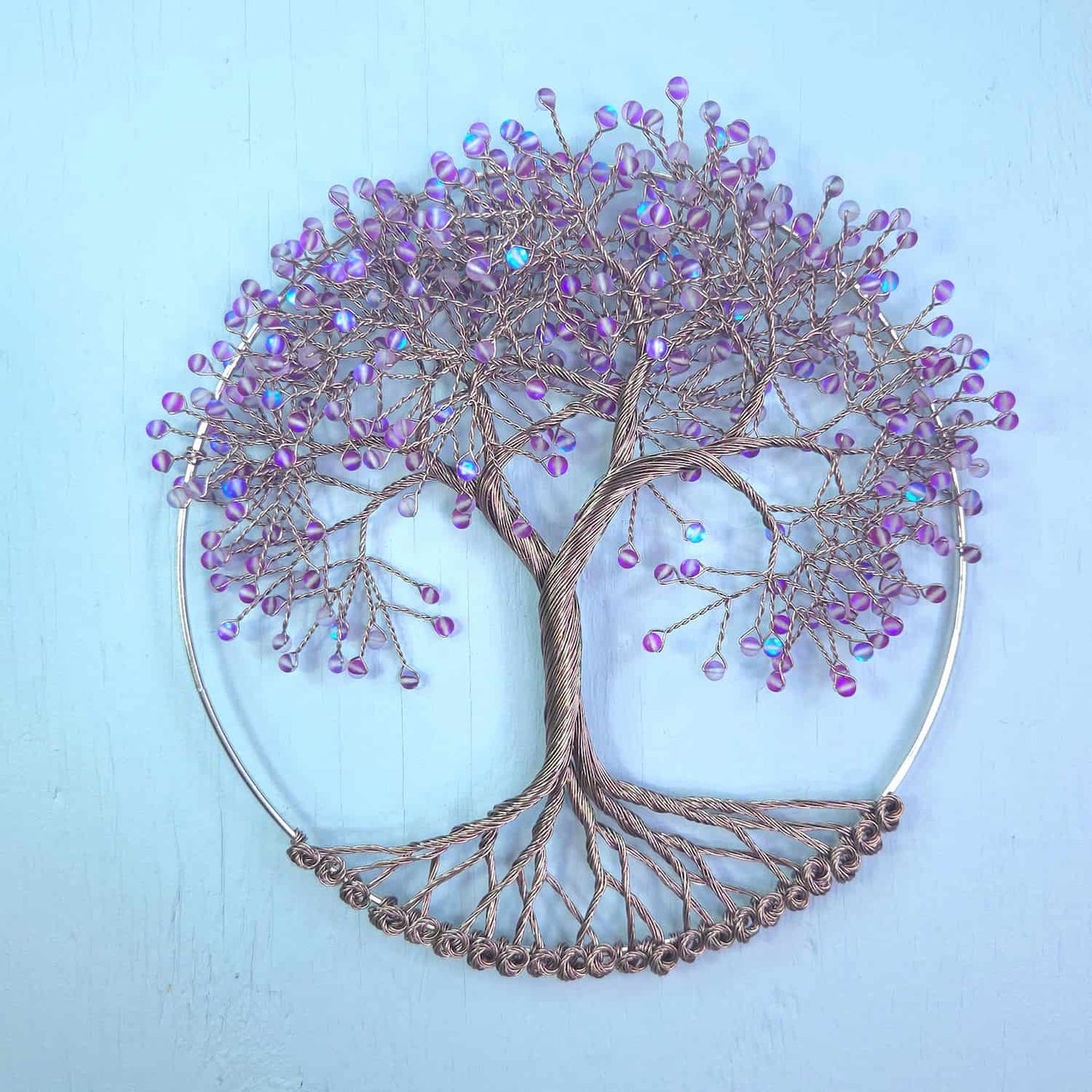 A circular wire tree of life wall art piece with a metallic bronze-brown trunk and branches, decorated with small iridescent purple beads, set against a light blue background.