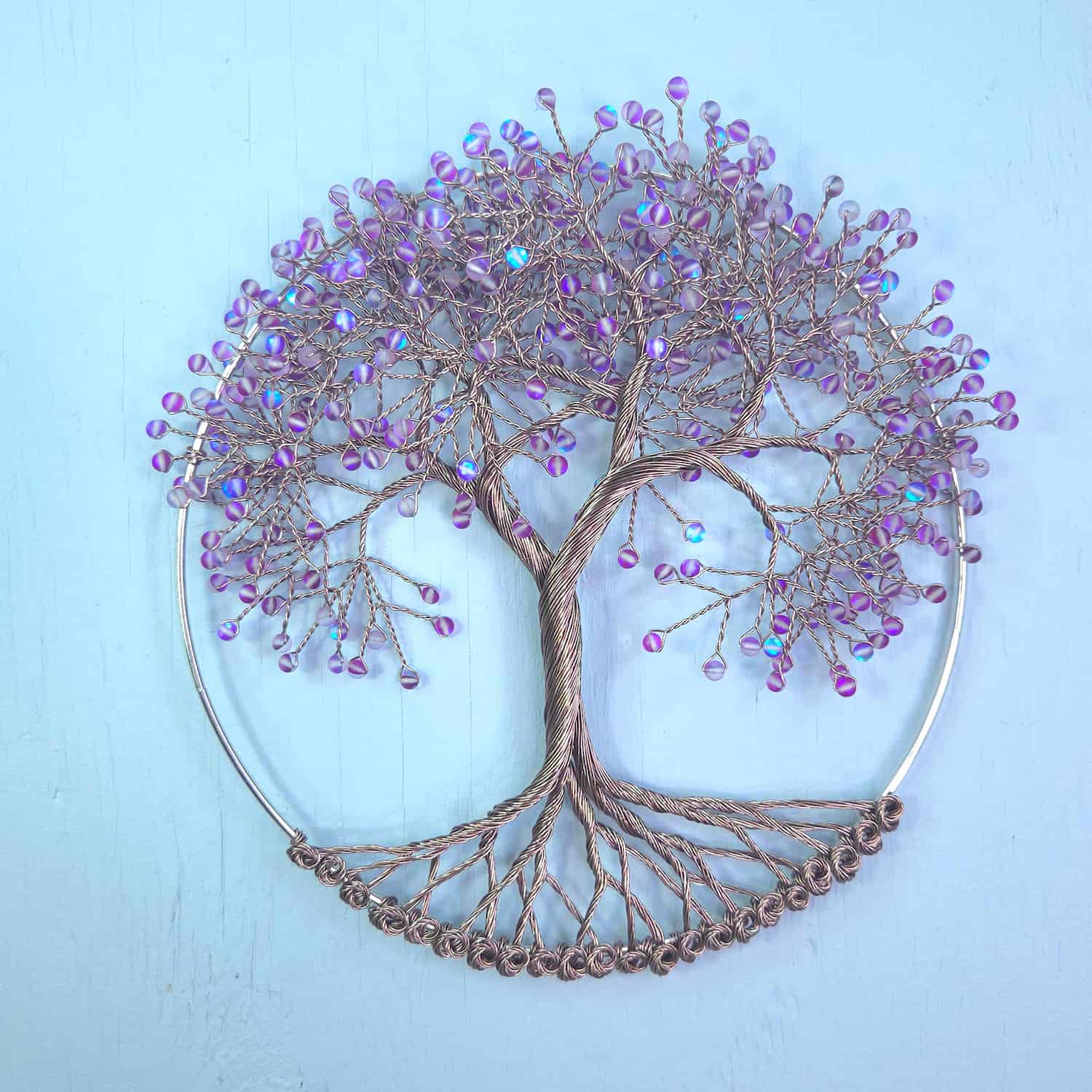 A circular wire tree of life wall art piece with a metallic bronze-brown trunk and branches, decorated with small iridescent purple beads, set against a light blue background.