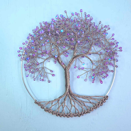 A circular wire tree of life wall art piece with a metallic bronze-brown trunk and branches, decorated with small iridescent purple beads, set against a light blue background.