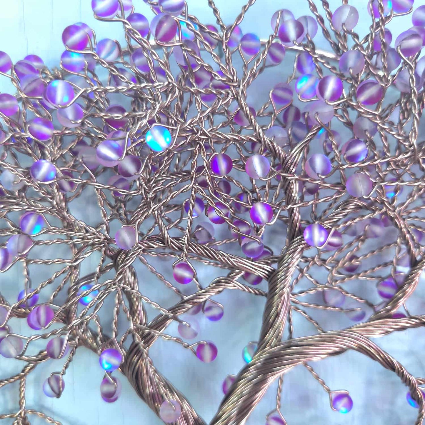 A decorative tree of life wire art piece with iridescent purple beads on a twisted copper wire trunk, set against a light blue background.