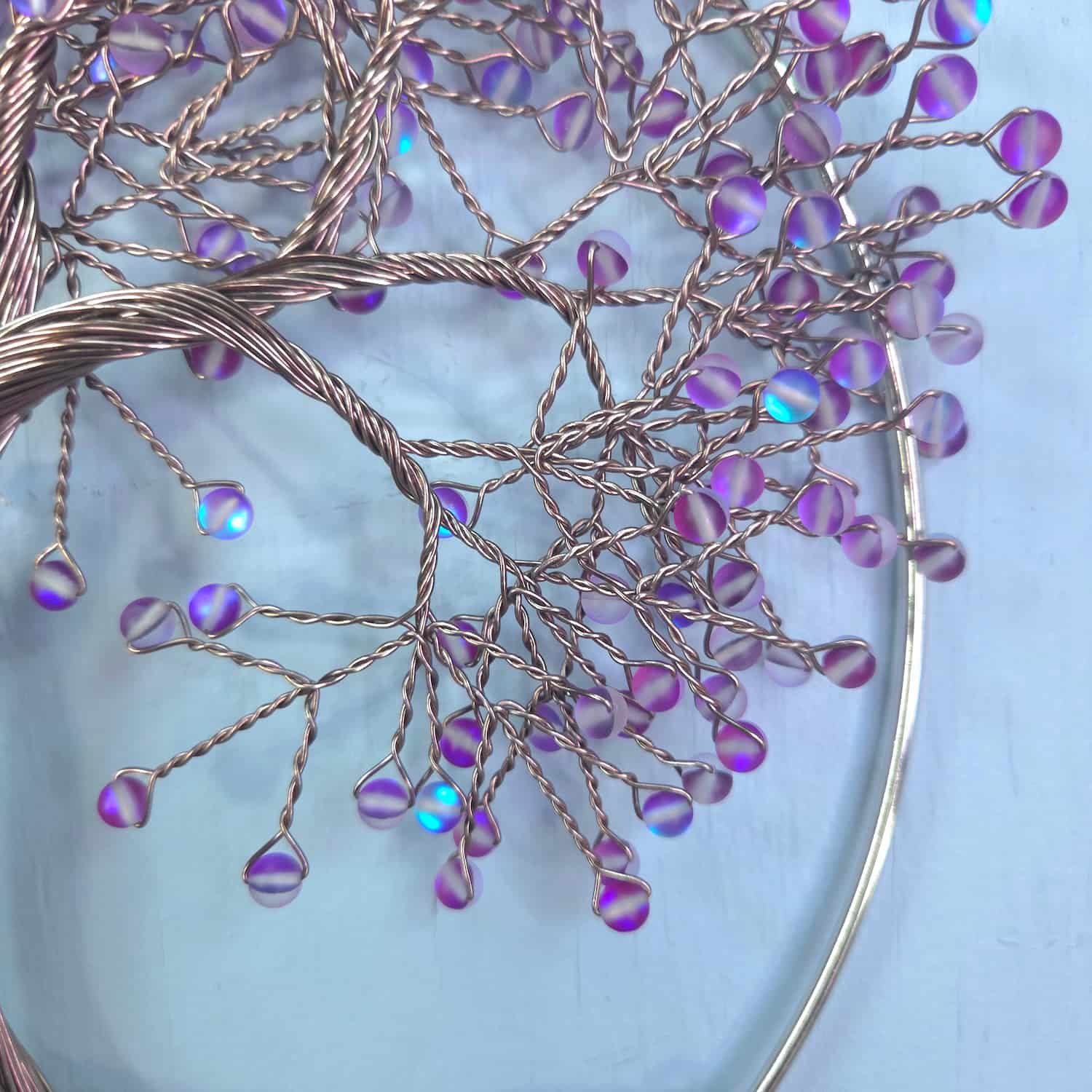 A close-up of a decorative wire tree of life ornament with iridescent purple and blue beads on copper-colored wire, displayed on a light blue background.