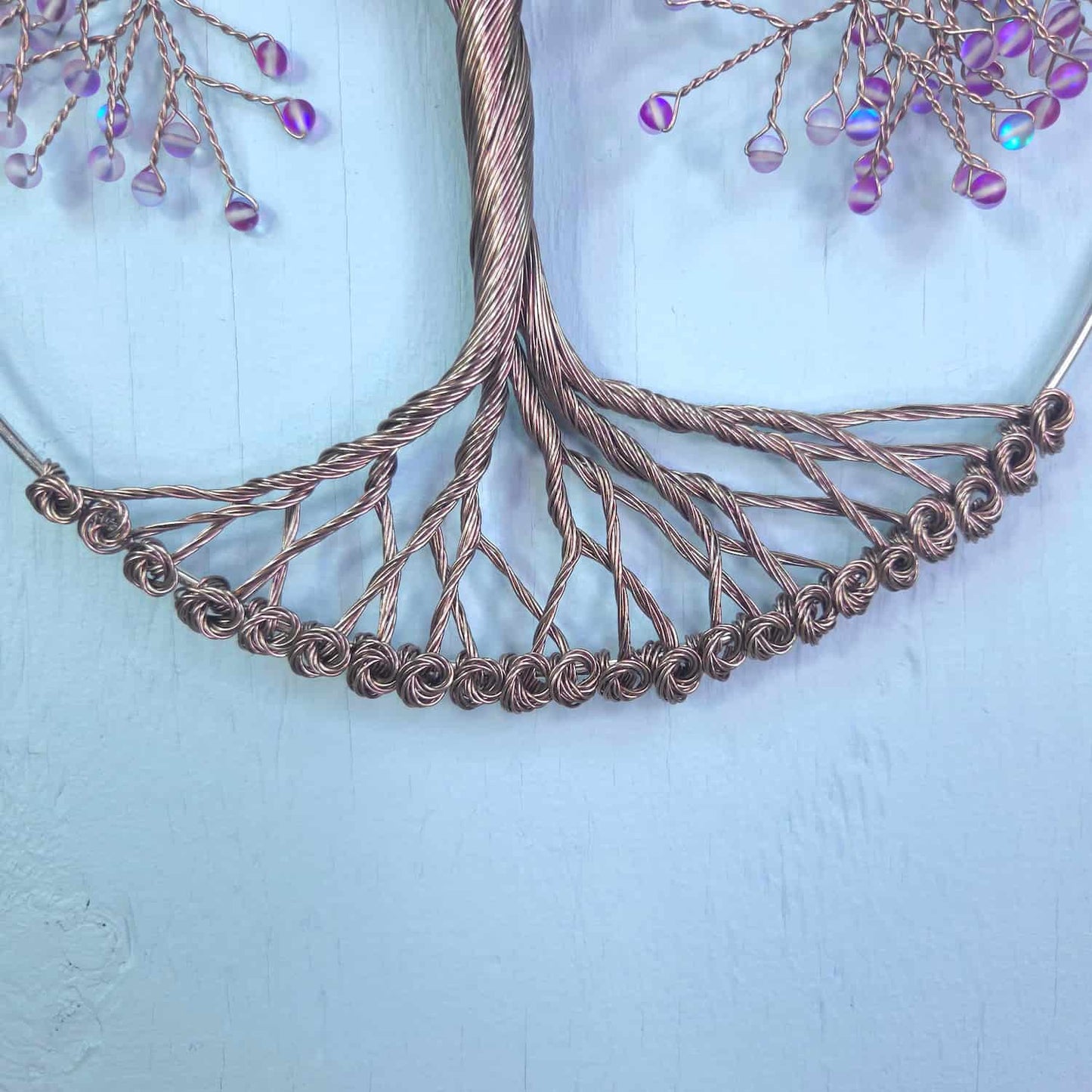 A copper-colored wire tree of life wall hanging with purple iridescent beads, displayed against a light blue painted wooden background.