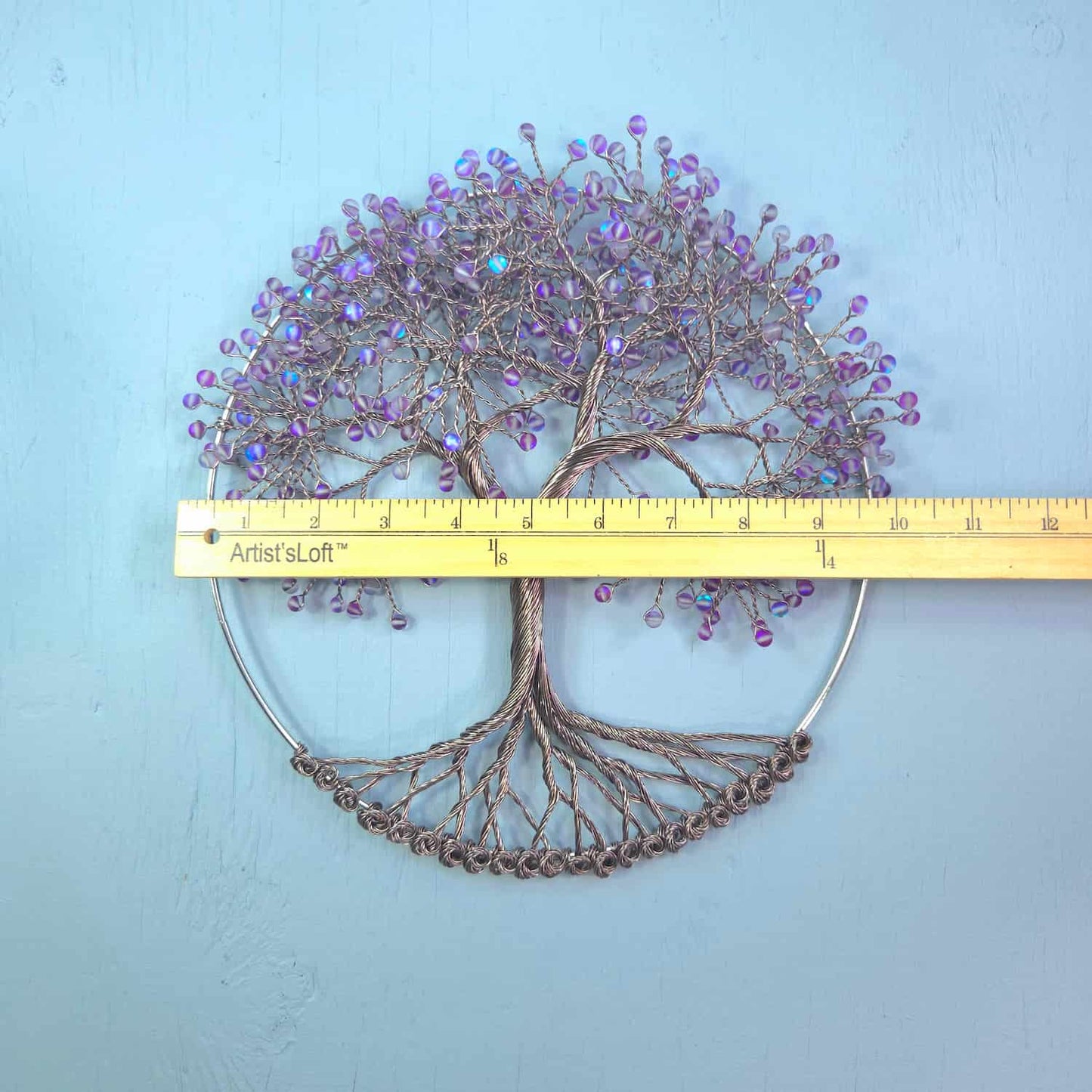 A decorative wire tree of life wall hanging with purple and blue iridescent beads, shown against a light blue background with a yellow Artist's Loft ruler across the middle for scale.