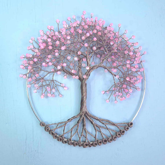A decorative metal wall art piece shaped like a tree with pink cherry blossoms, mounted on a circular silver hoop against a light blue background.