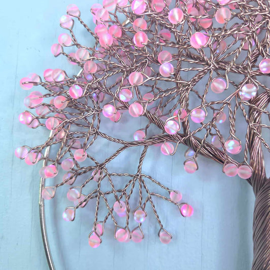 A close-up of a decorative tree branch sculpture with pink iridescent bead-like droplets on copper-toned wire branches, set against a light blue background.