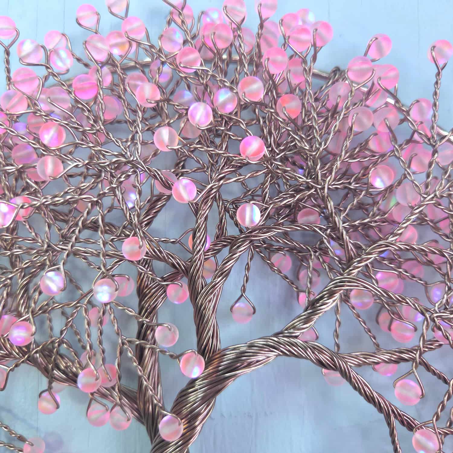 A decorative wire tree of life wall art piece with pink iridescent bead blossoms on twisted metallic branches, shown against a light blue background.