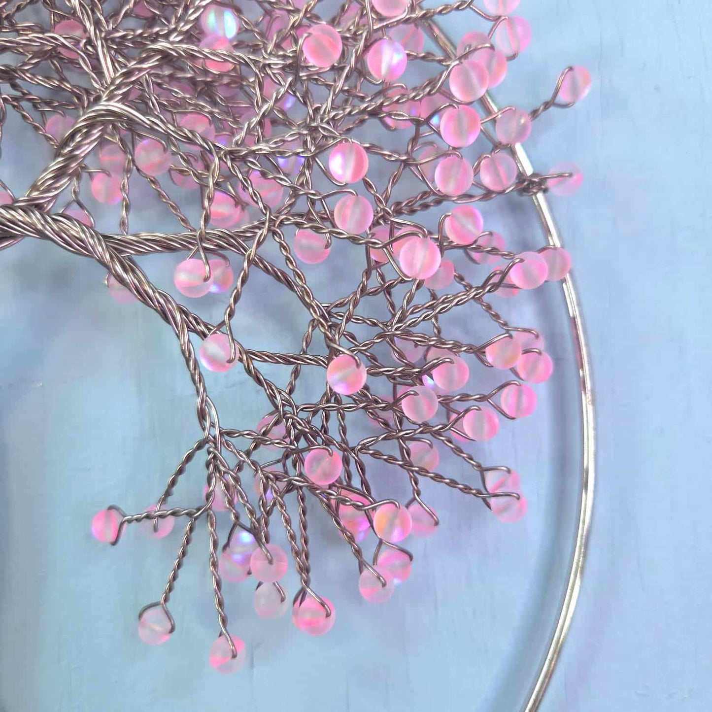 A close-up of a decorative branch-like wire sculpture with many small pink iridescent bead-like droplets, arranged on a pale blue background.