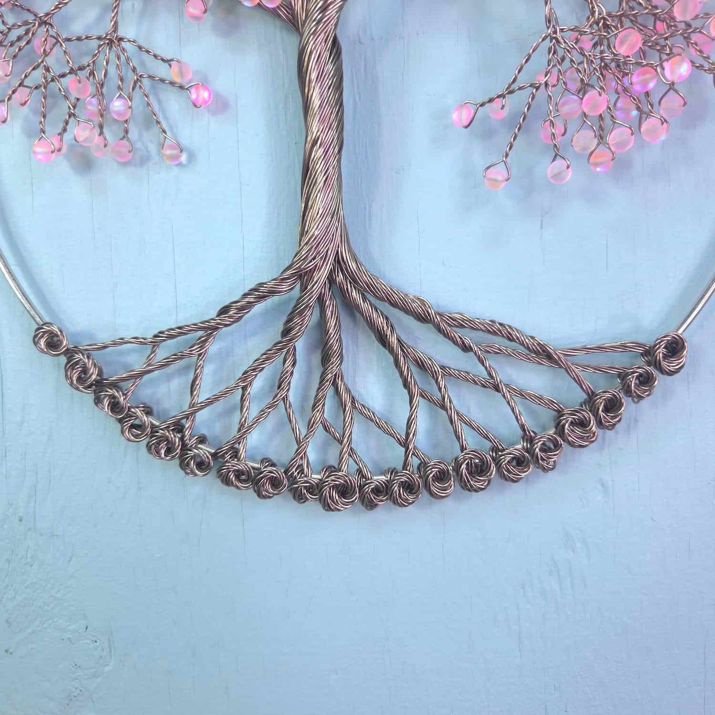 A close-up of a metallic wire tree branch or wreath design with twisted copper-toned branches and pink faceted bead accents against a light blue background.