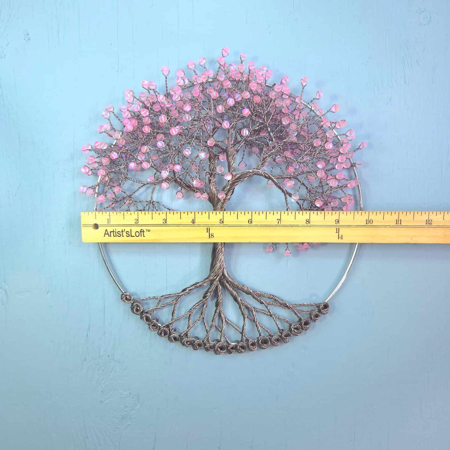 A wire art wall sculpture of a tree with pink cherry blossoms and exposed roots, displayed against a light blue background with a yellow ArtistLoft brand tape measure placed horizontally across the middle for scale.