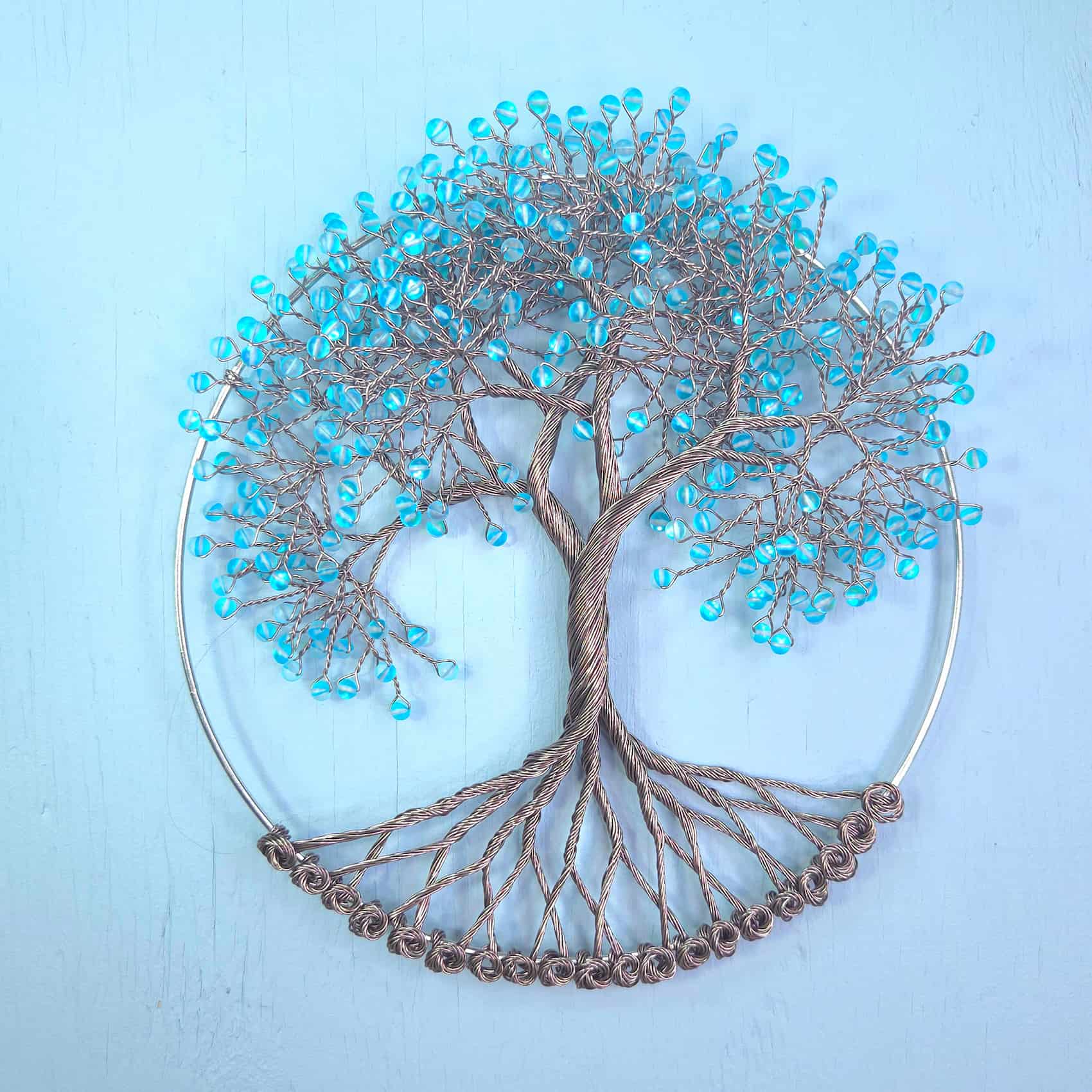 Wire tree of life wall art with turquoise blue seed beads on branches, mounted on a circular metal hoop against a light blue background.
