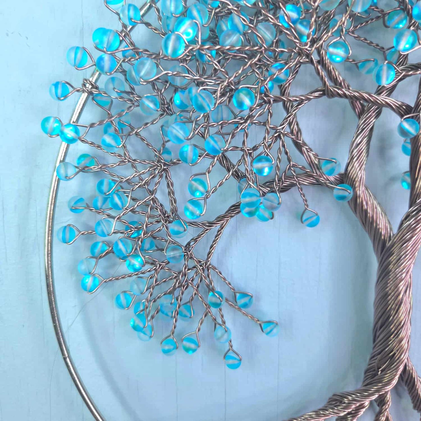 A close-up of a decorative wire tree of life wall hanging with metallic silver branches and translucent light blue beads, set against a pale blue background.