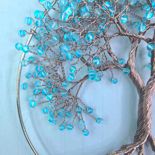A close-up of a decorative wire tree of life wall hanging with metallic silver branches and translucent light blue beads, set against a pale blue background.
