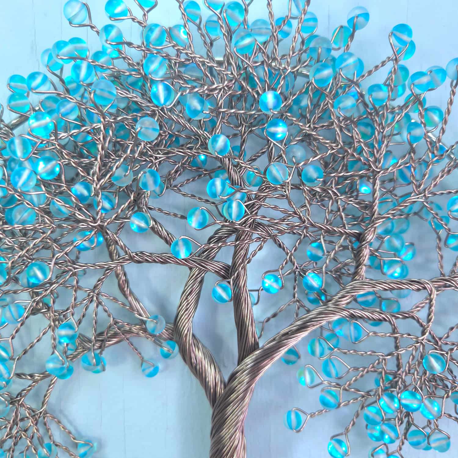 A decorative metal tree of life wall hanging with twisted silver branches and numerous round, light blue glass beads forming the foliage, set against a light blue background.