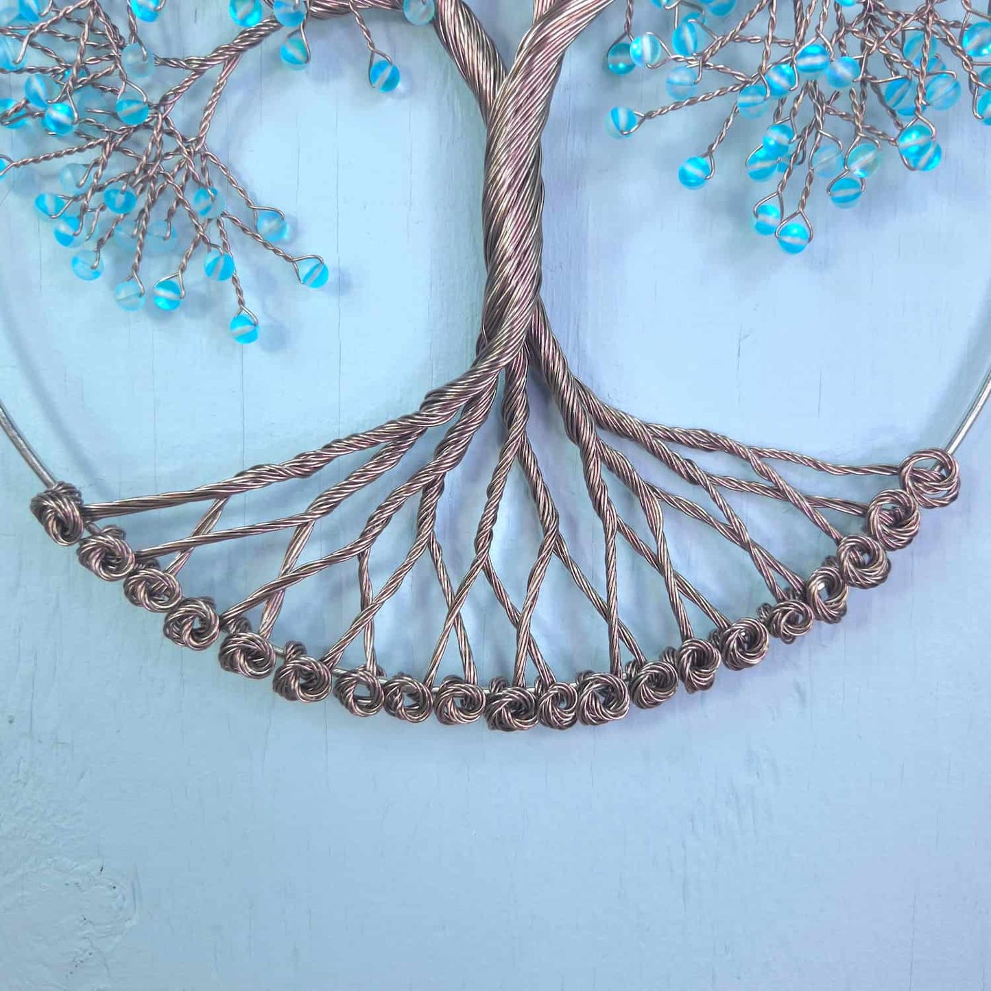 A decorative wire tree of life wall hanging with a copper-toned twisted trunk and branches, embellished with small light blue faceted beads, shown against a pale blue textured background.