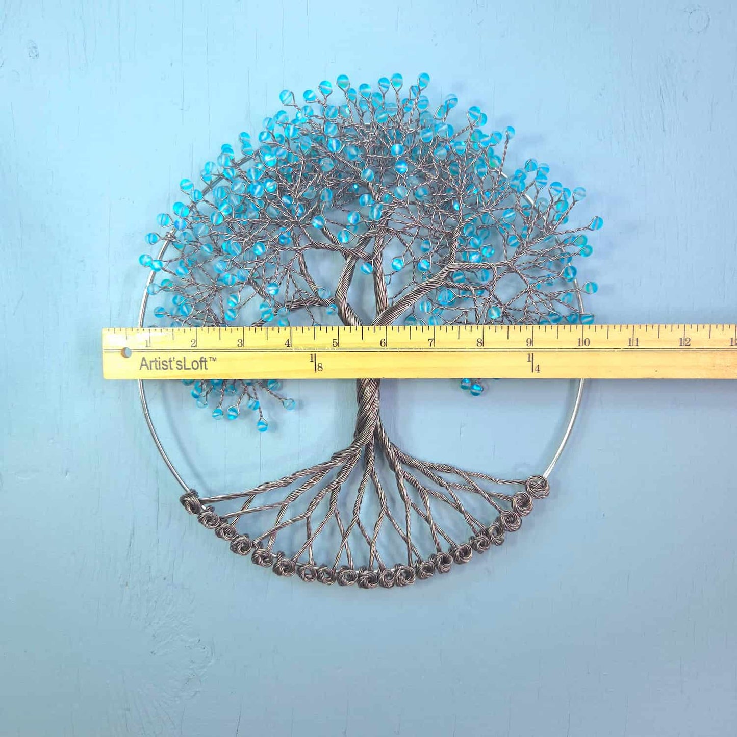 A wire tree-of-life wall hanging with turquoise blue beads and a circular metal frame, shown with a yellow measuring tape across the middle to indicate scale against a light blue textured background.