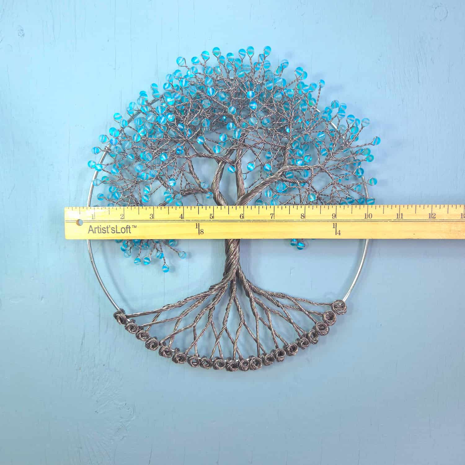 A wire tree-of-life wall hanging with turquoise blue beads and a circular metal frame, shown with a yellow measuring tape across the middle to indicate scale against a light blue textured background.